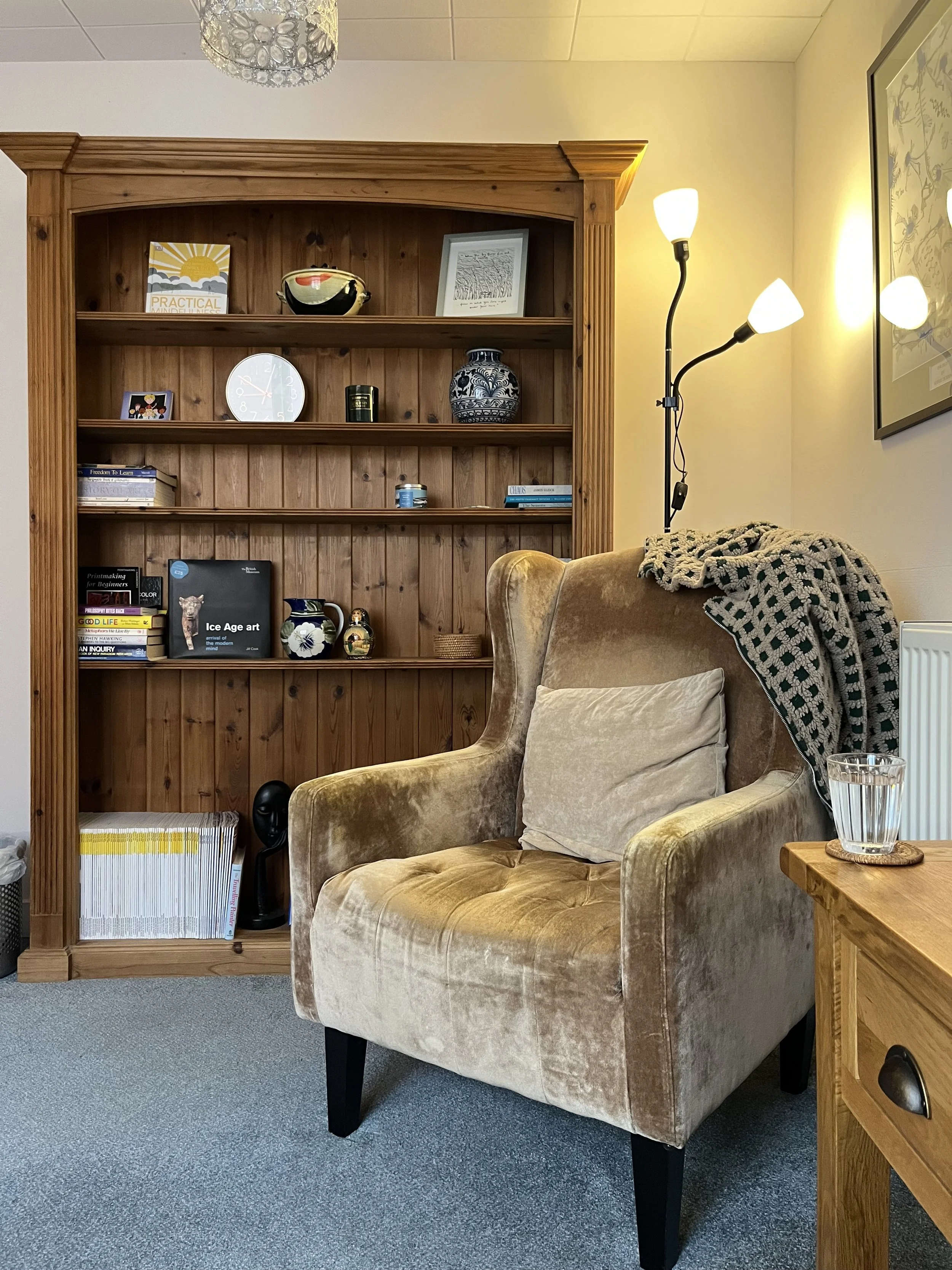 A cozy living room corner with a beige velvet armchair, a wooden side table with a glass of water, a bookshelf with books and decorative items, a wooden bookcase, and a tall floor lamp with three adjustable white lampshades. There is also a framed picture on the wall to the right.
