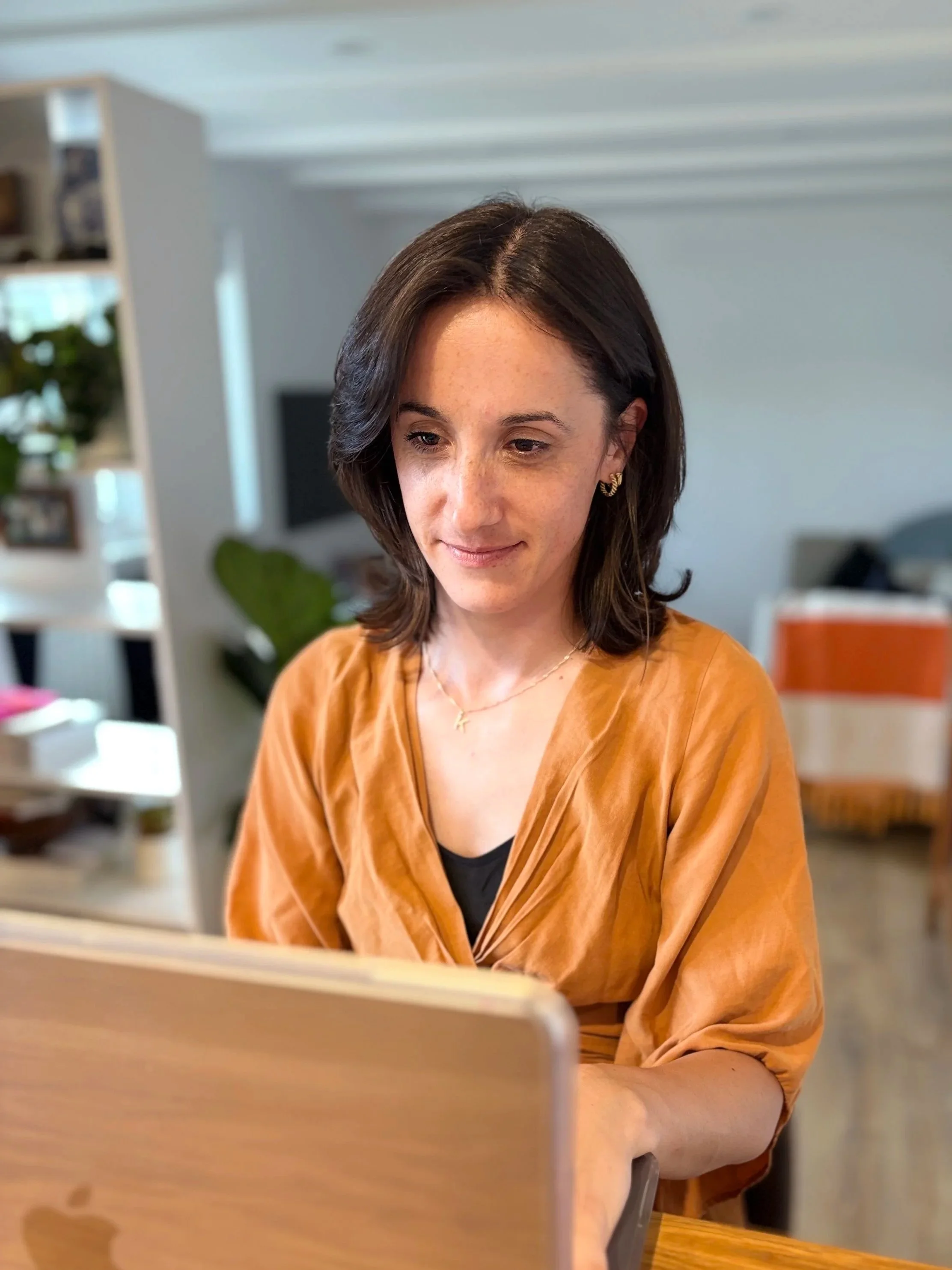 A woman with shoulder-length dark brown hair, wearing a black top and an orange-brown wrap shirt, sitting at a desk using a laptop.