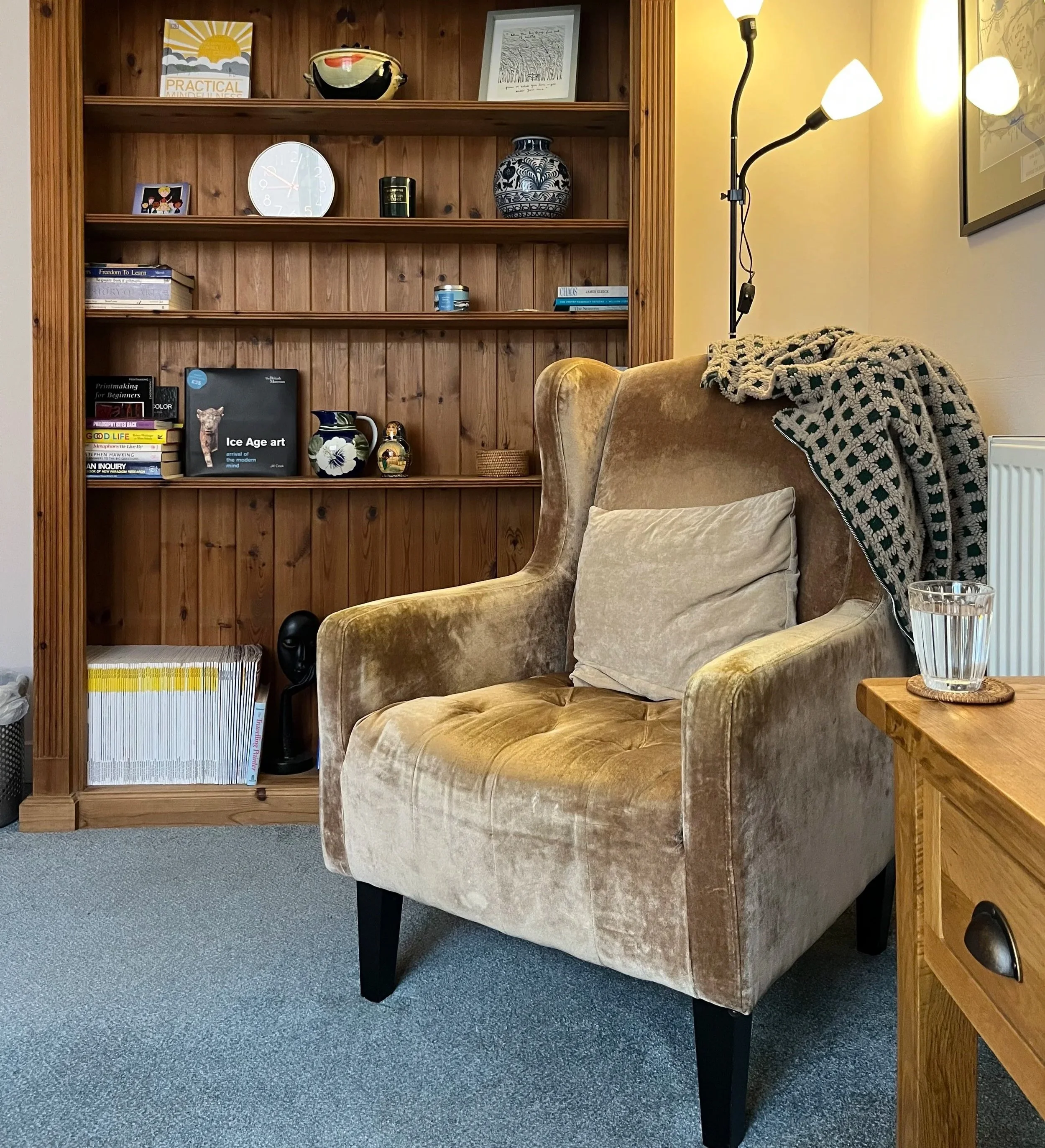 View of a cozy counselling room with a beige armchair, a small wooden side table with a glass of water, and a tall wooden bookshelf filled with books and decorative items, illuminated by a floor lamp.