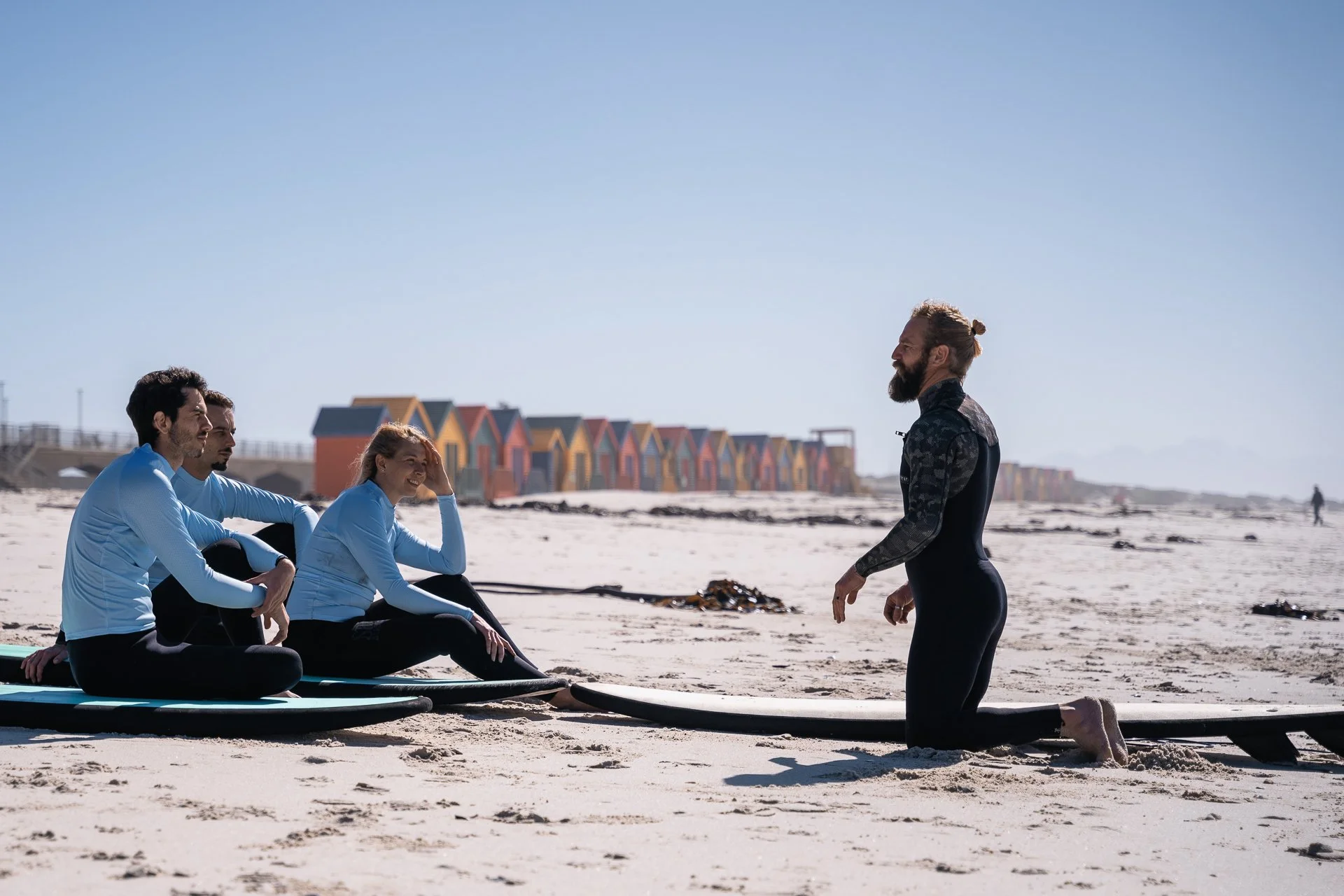 A surf instructor coaches a small group of students sitting on the sand, with colourful Muizenberg beach huts in the background.