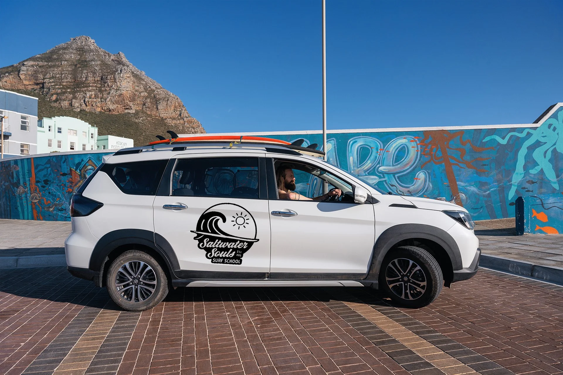 Saltwater Souls branded car with a surfboard on the roof, parked in front of a colourful street mural with Lion's Head mountain behind.