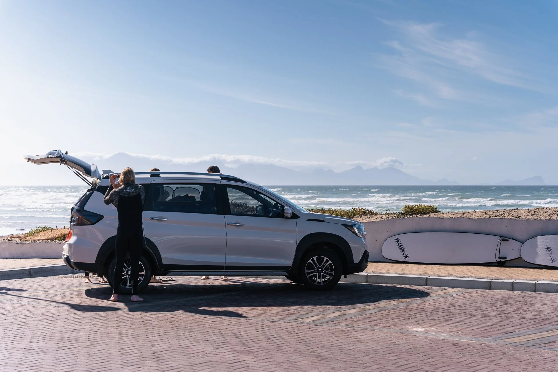 A woman loads a surfboard into the boot of a car parked on the beachfront, with more surfboards nearby and the ocean and mountains behind.