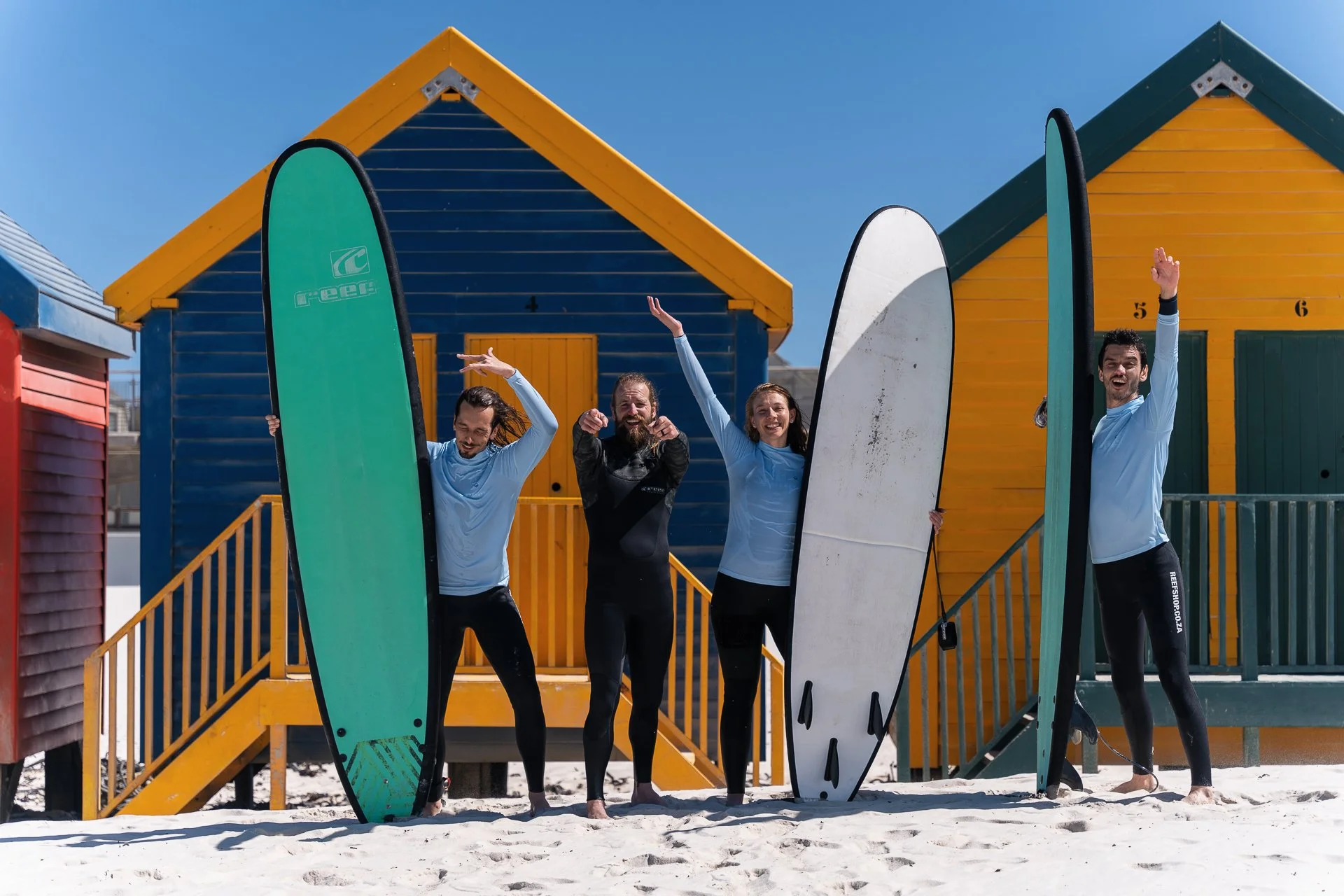 Four surfers cheering and holding surfboards aloft in front of the colourful Muizenberg beach huts.