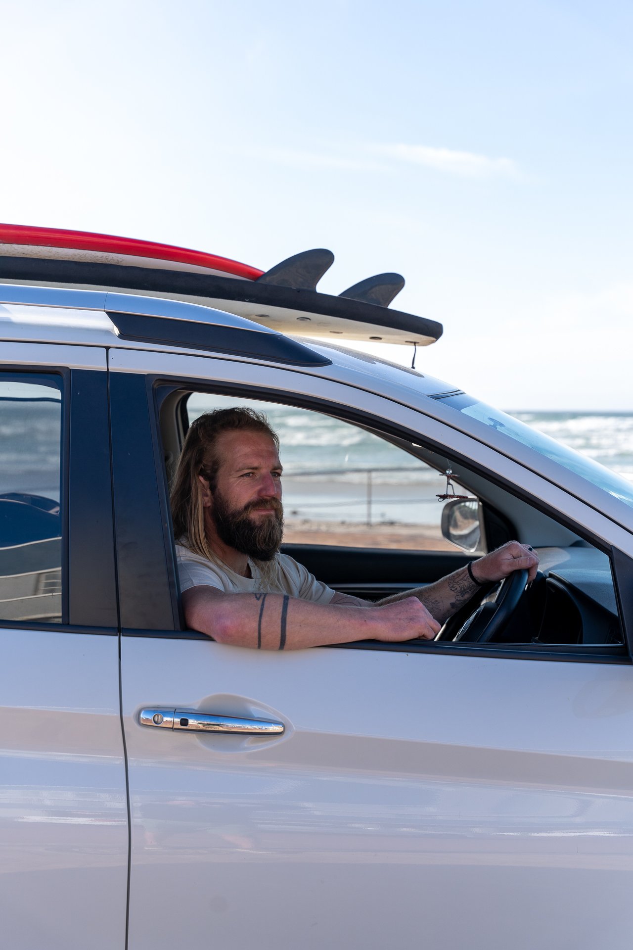 A bearded man sits in the driver's seat of a car with a surfboard strapped to the roof rack, parked near the beach.