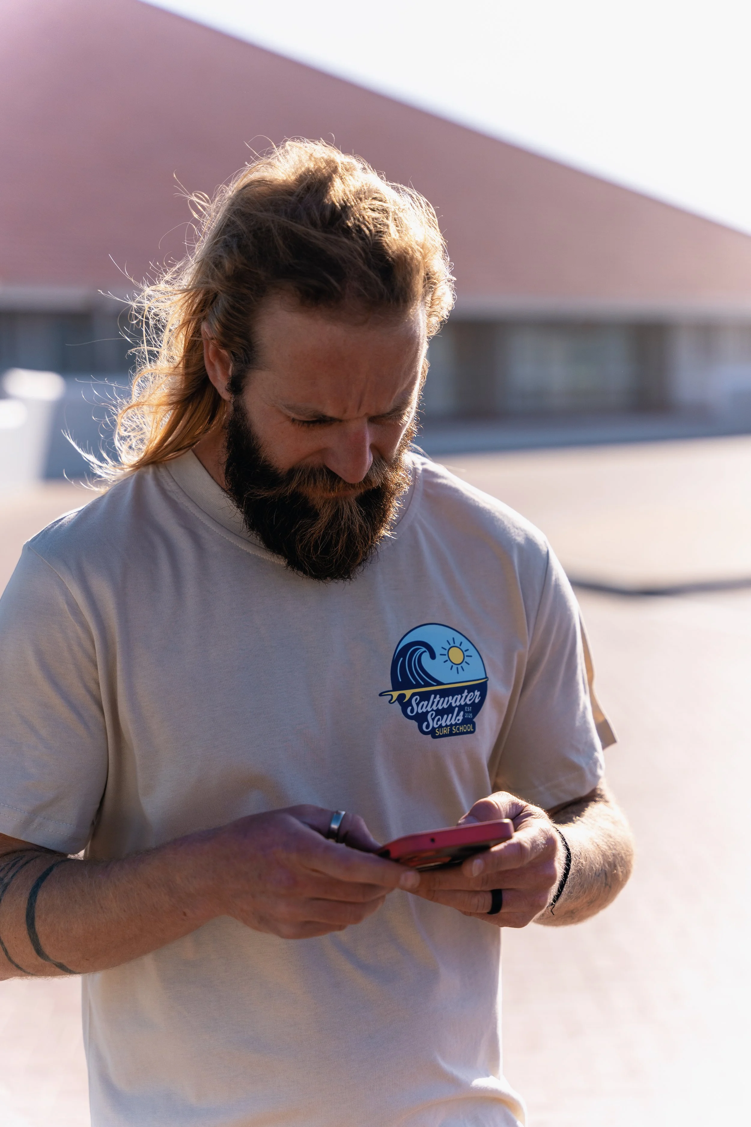 Henri from Saltwater Souls Surf School, wearing the brand t-shirt, looking down thoughtfully outdoors.