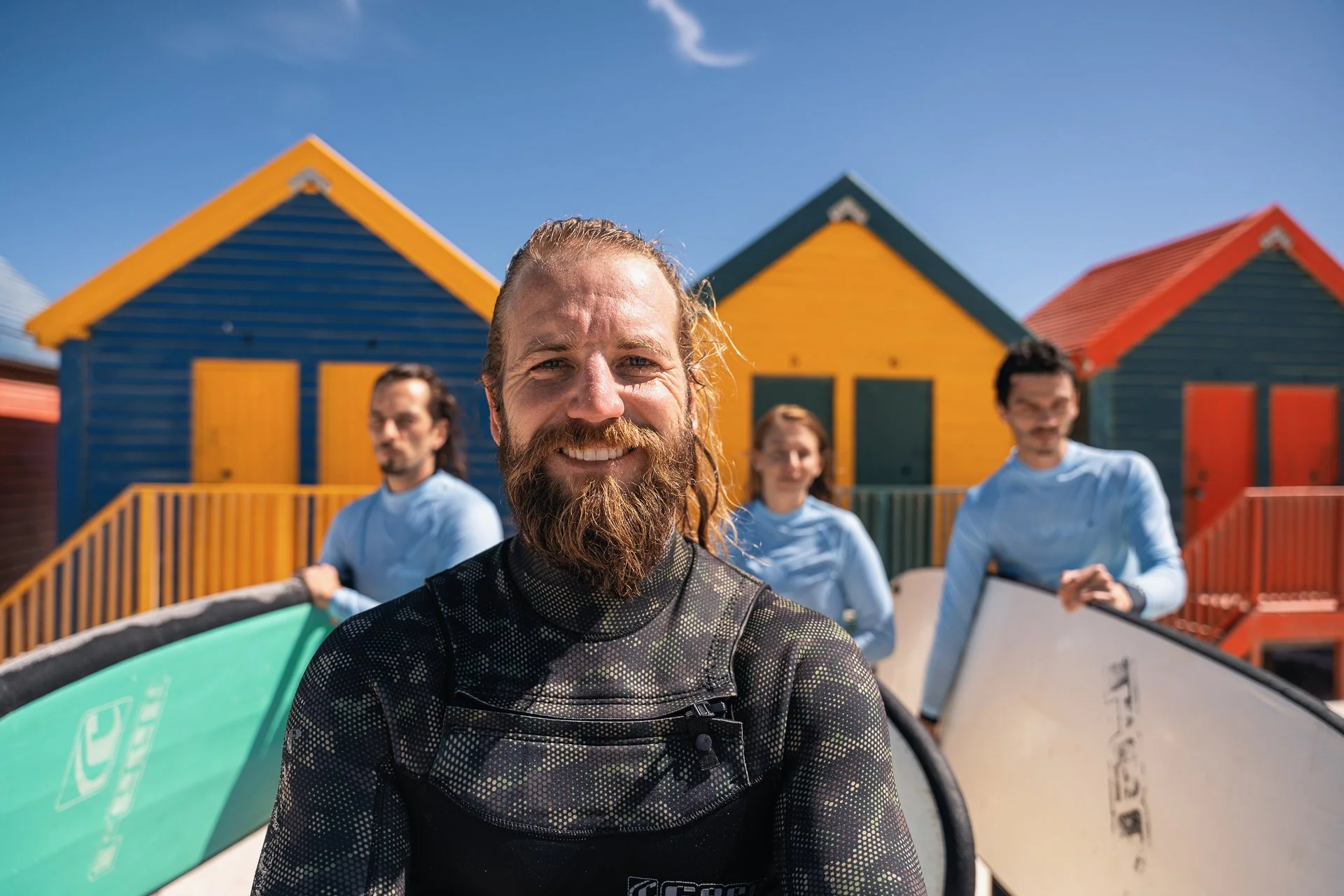 Henri, founder and lead instructor of Saltwater Souls, smiling with fellow surfers in wetsuits in front of colourful beach huts.