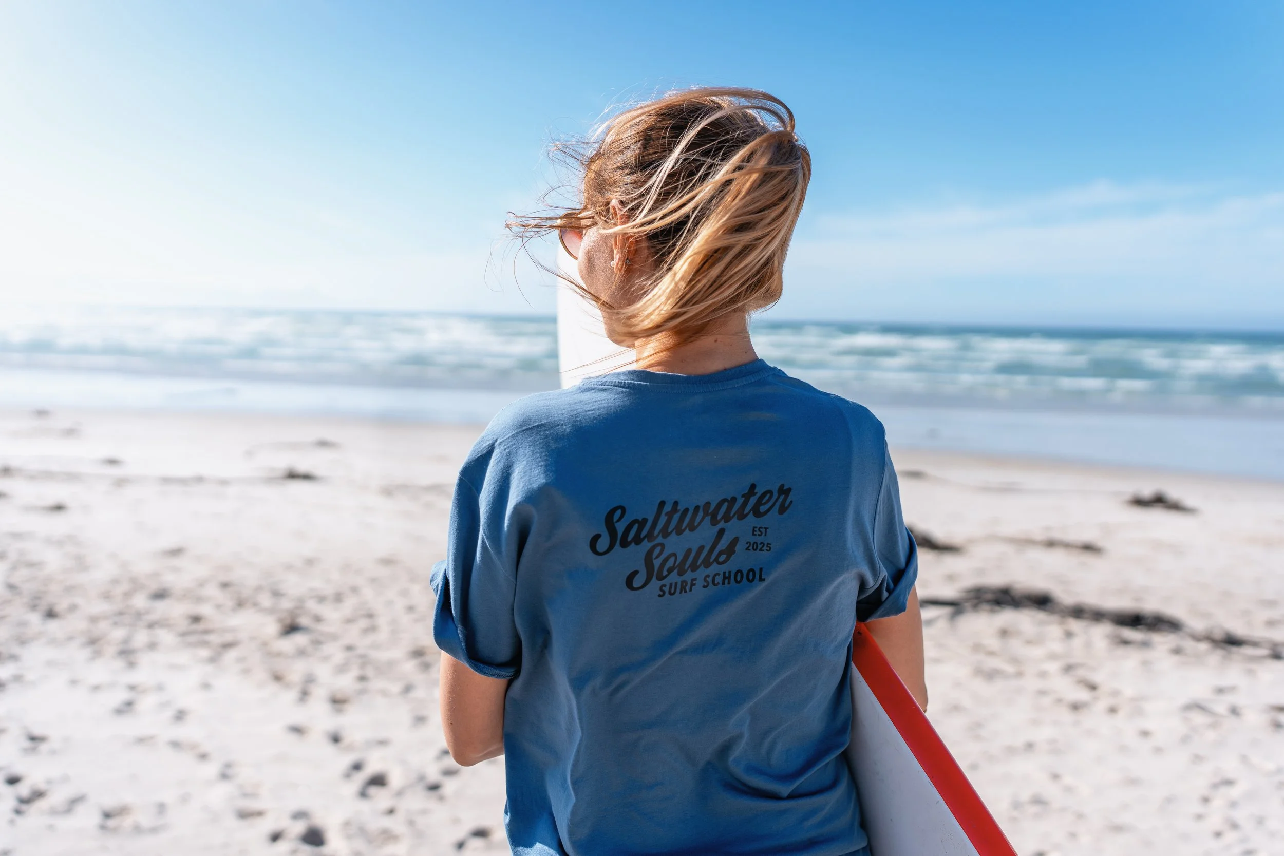 Woman with windswept hair wearing a Saltwater Souls Surf School t-shirt, carrying a surfboard on a Cape Town beach.
