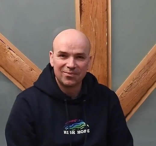 A bald man wearing a black hoodie with a rainbow graphic and the words 'High Hope' sitting indoors, with wooden beams in the background.