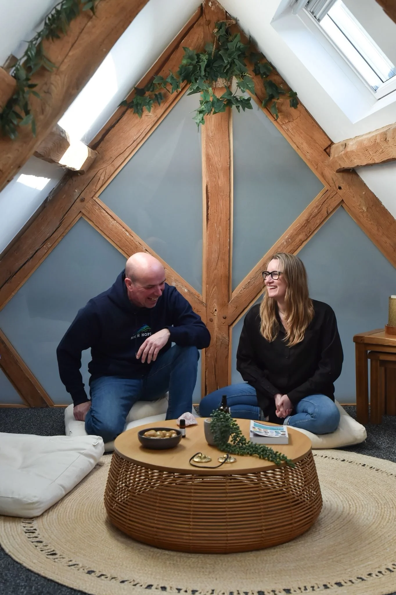 A man and woman sitting on cushions and laughing around a round wooden coffee table in a cozy attic space with wooden beams and blue wall panels.