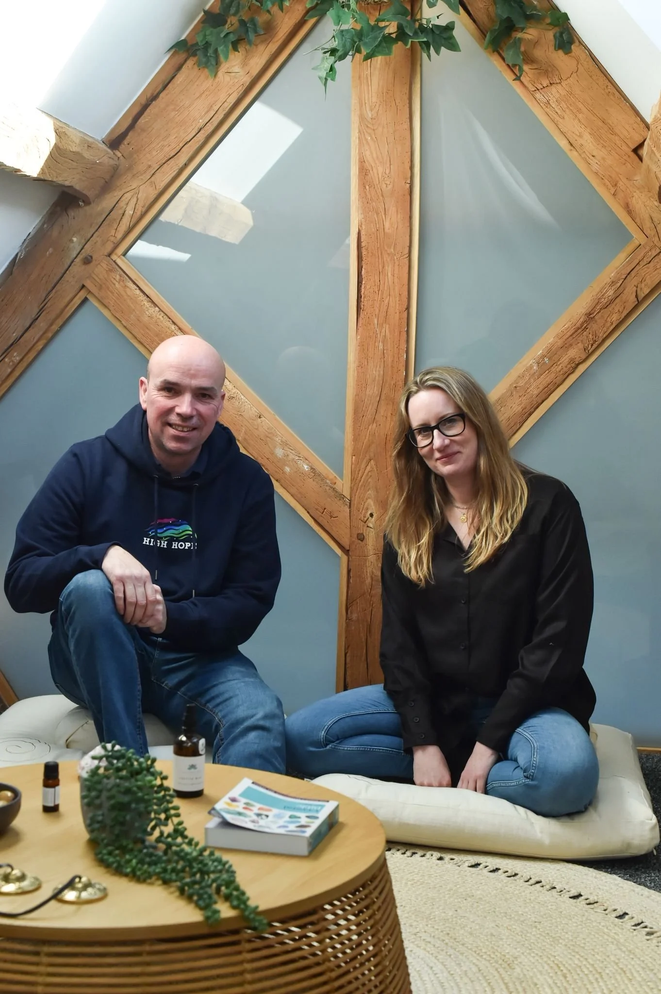 A man and a woman sitting on a cushion in front of a wall with wooden beams and frosted glass panels. The table in front has a small plant, bottles of essential oils, and a book.