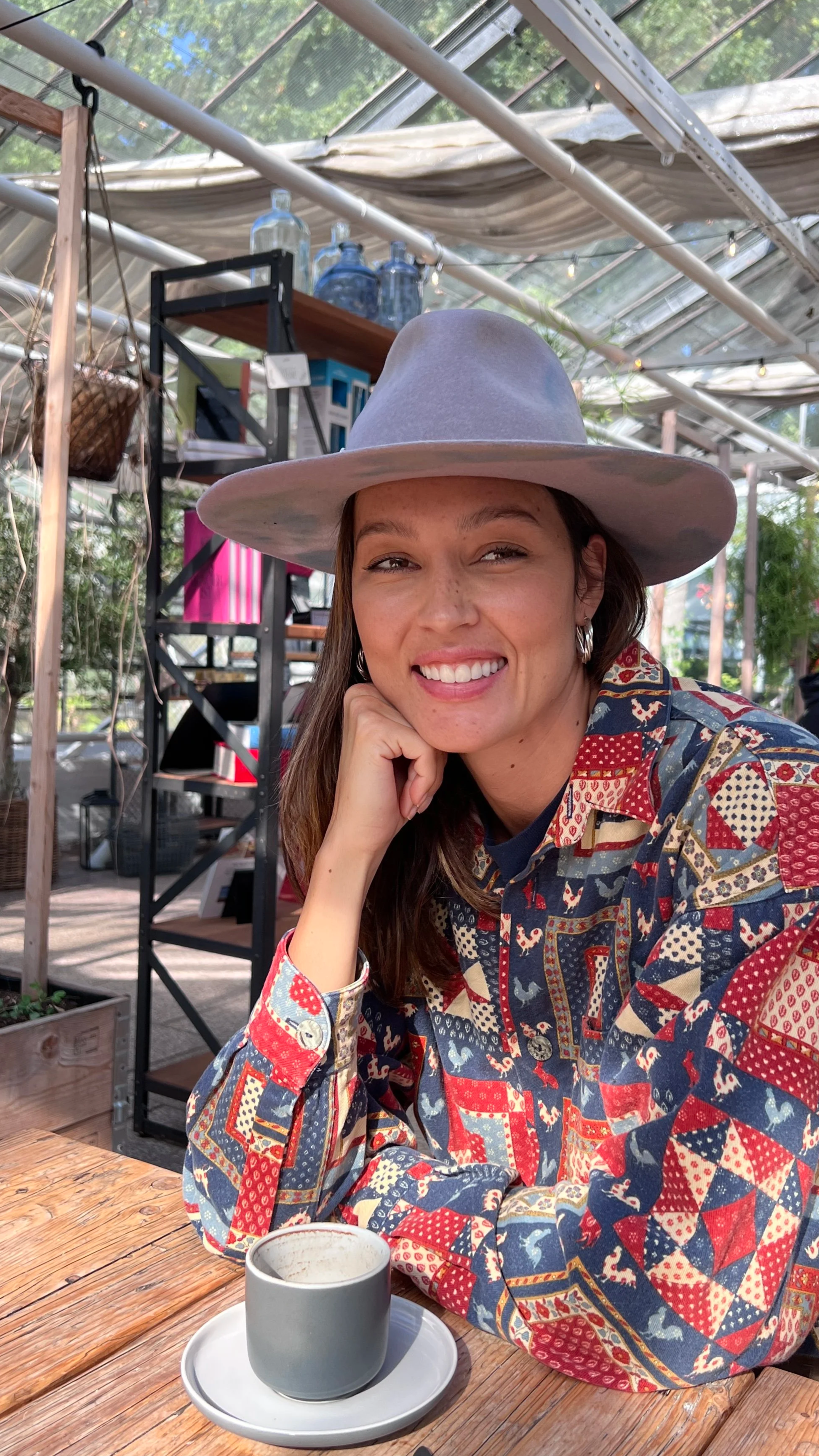 Junge Frau trägt einen handgefertigten grauen Fedora-Hut von Hut Atelier. Natürliches Licht, Holzdetails und Pflanzen betonen den Boho-Stil und die nachhaltige Ästhetik der Marke.