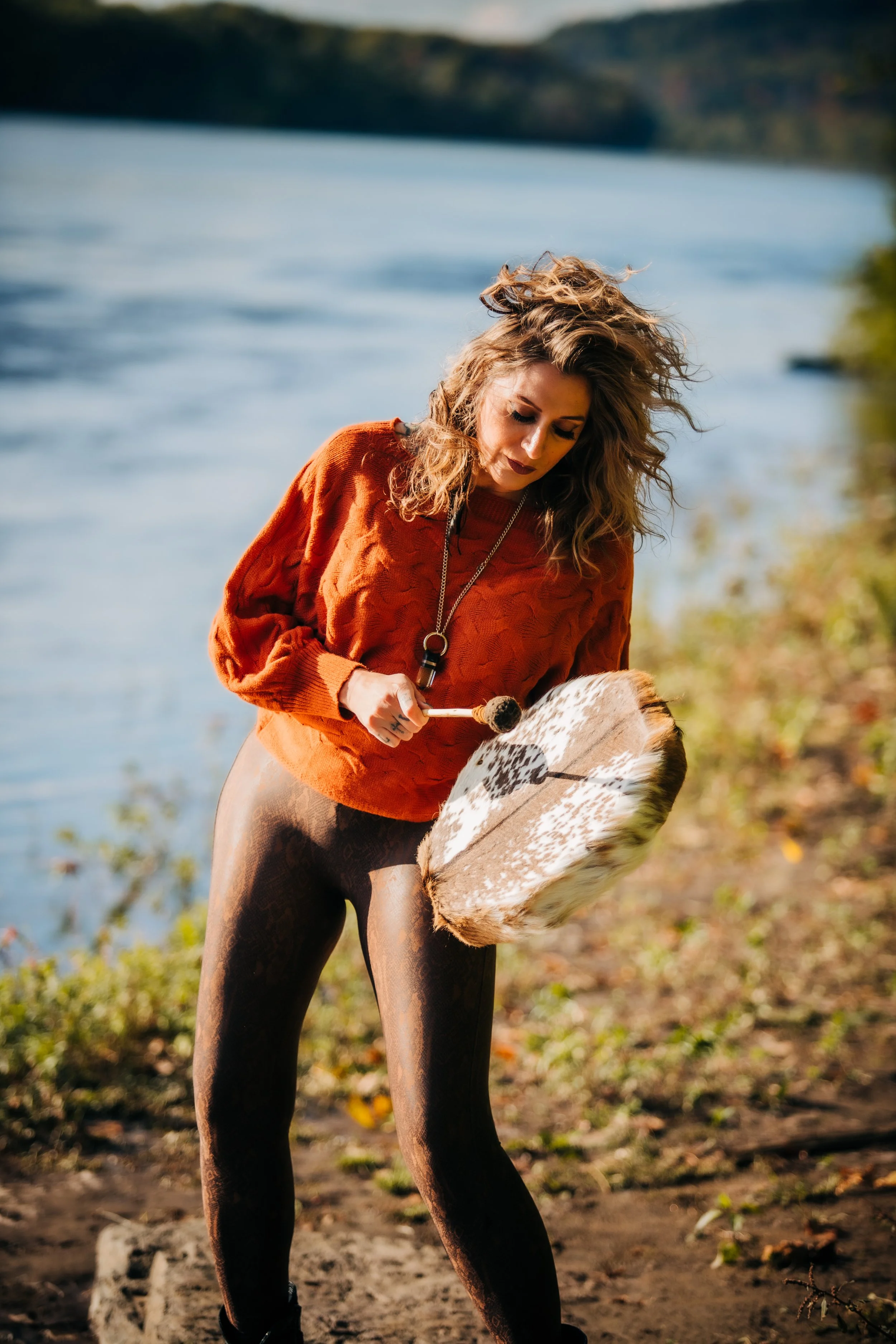 A woman with curly hair wearing a red sweater and patterned leggings playing a drum by a river.