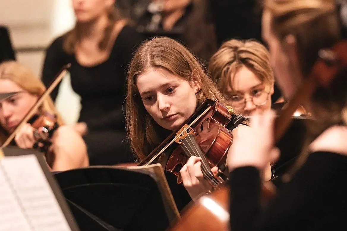 Young girl playing the violin during a symphony rehearsal, surrounded by other musicians.