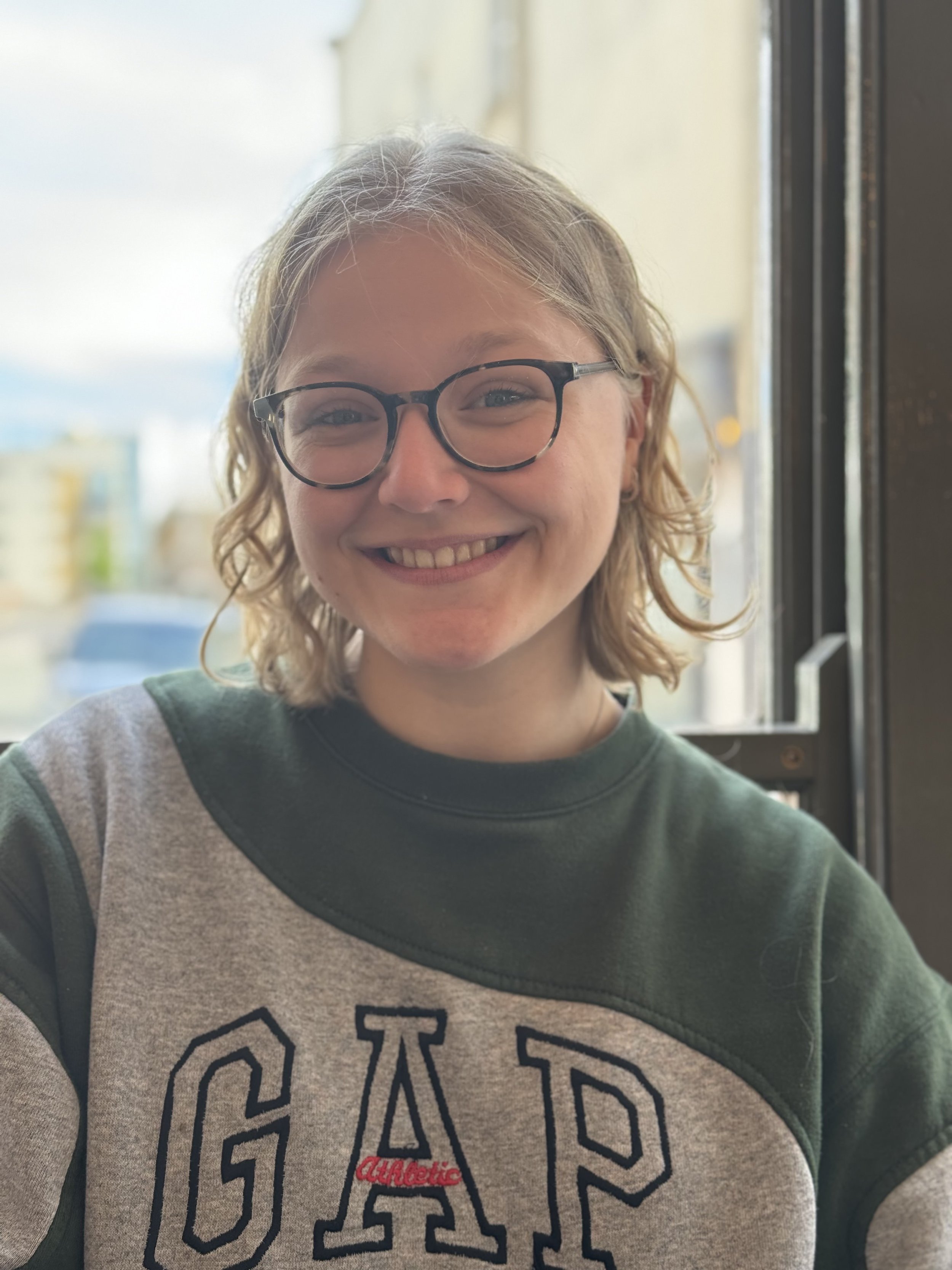 A young woman with shoulder-length curly blond hair, glasses, and a big smile, sitting near a window with city buildings seen in the background. She is wearing a green and gray sweatshirt with the word 'GAP' on it.