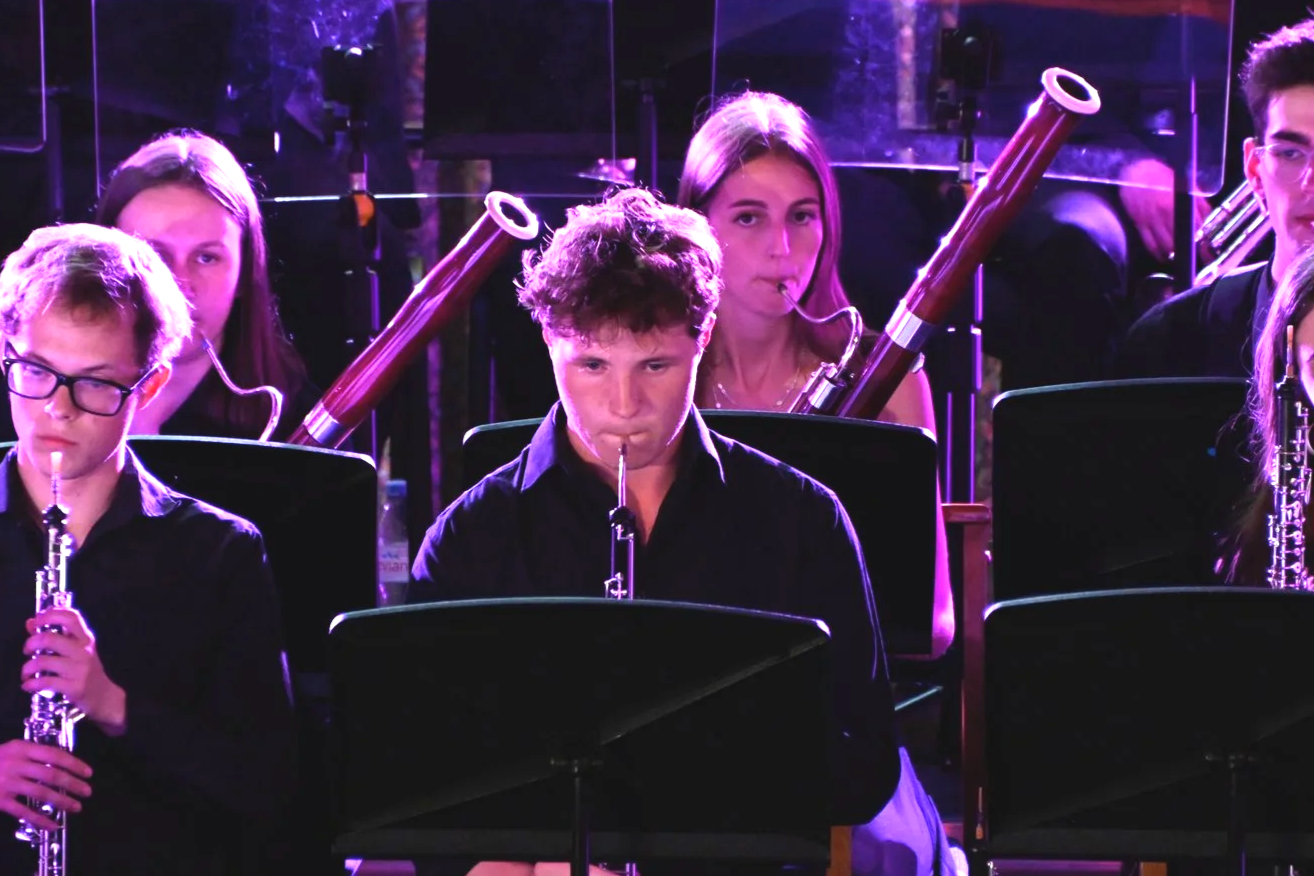 Young musicians in an orchestra play wind instruments, including clarinets and bassoons, during a performance under purple lighting.