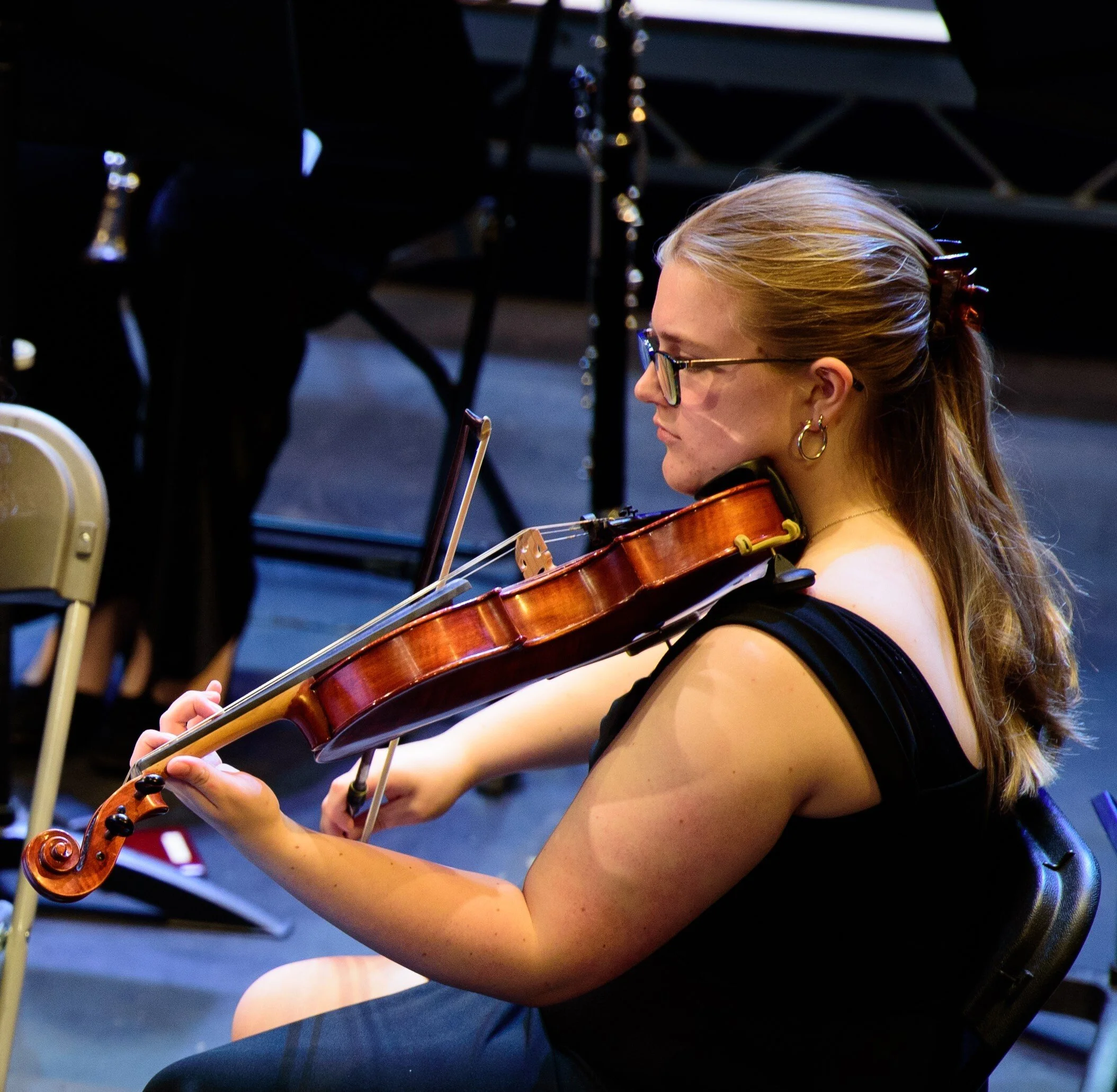 A woman with blonde hair, wearing glasses and a black dress, playing a violin.