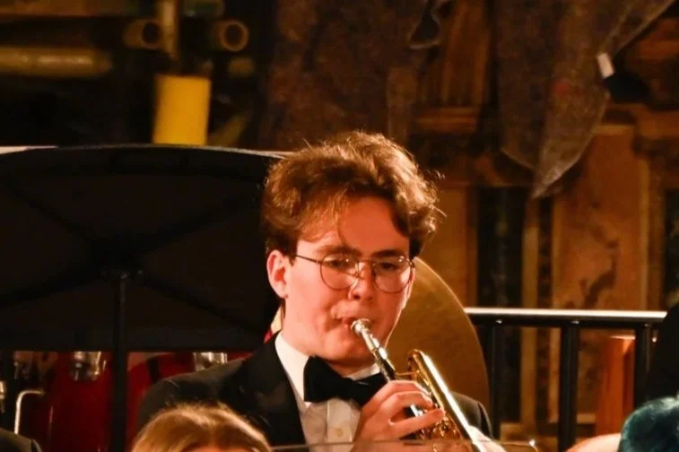 Young man in tuxedo with glasses playing a trumpet in an indoor concert setting with warm lighting.