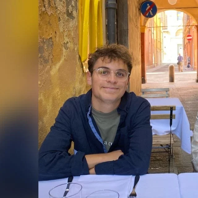 Young man with glasses sitting at a table outdoors in a colorful European street, smiling with arms crossed, with empty tables and chairs around him.