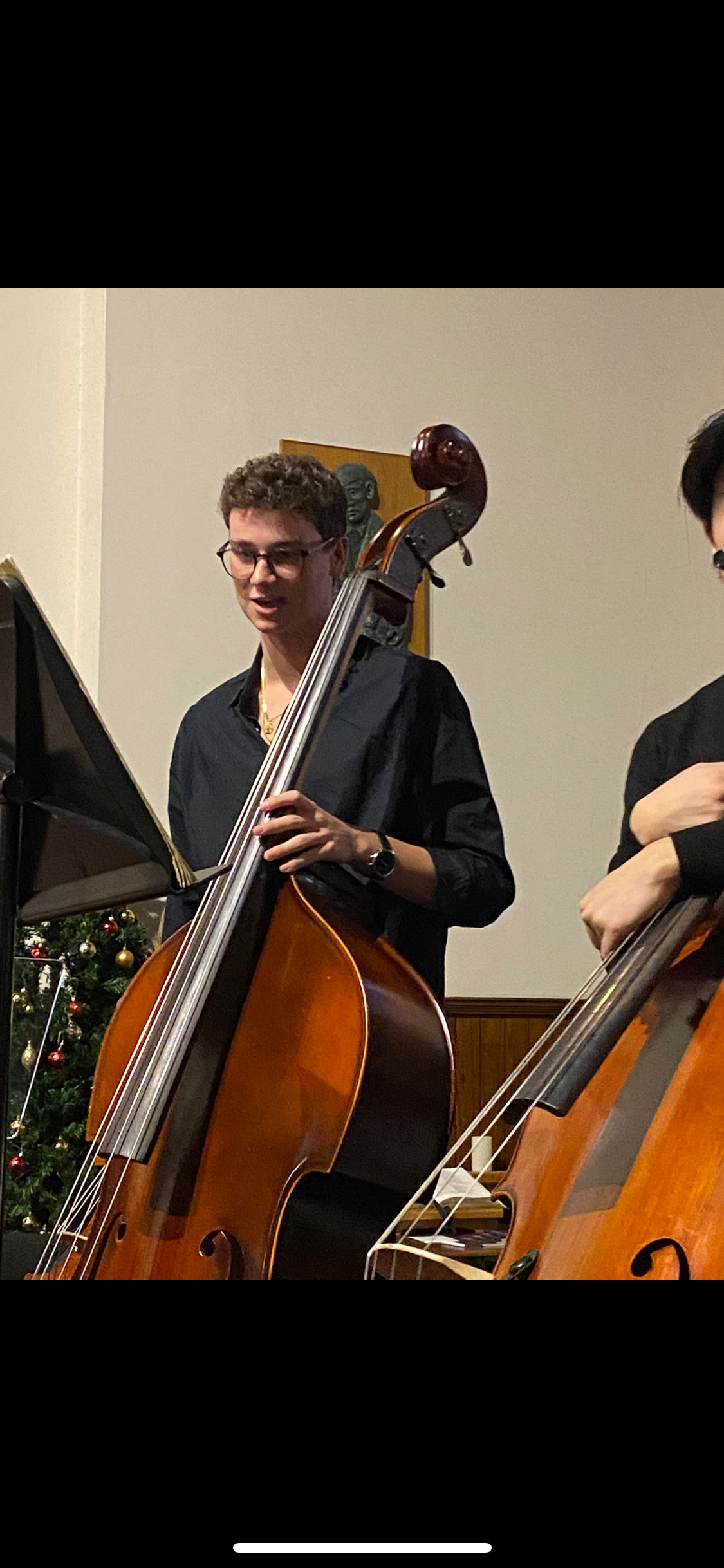 A young man with glasses playing an upright bass during a performance, with a Christmas tree and a small sculpture on the wall in the background.