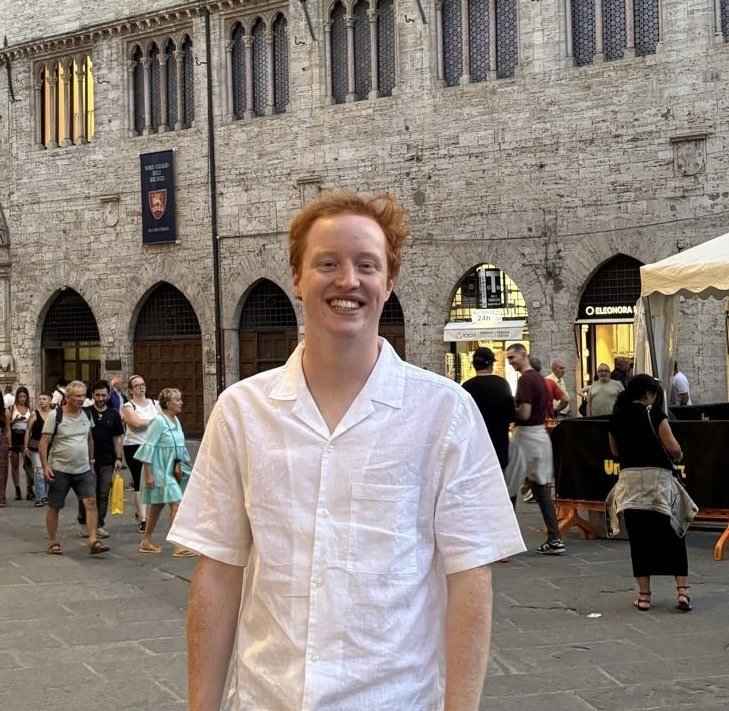 A young man with red hair smiling and wearing a white button-up shirt, standing in a busy square with historic stone buildings and people walking in the background.