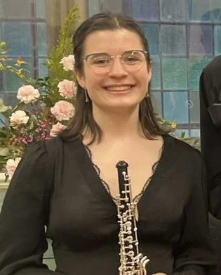 Woman with glasses holding a clarinet, smiling, with pink and white flowers in the background.