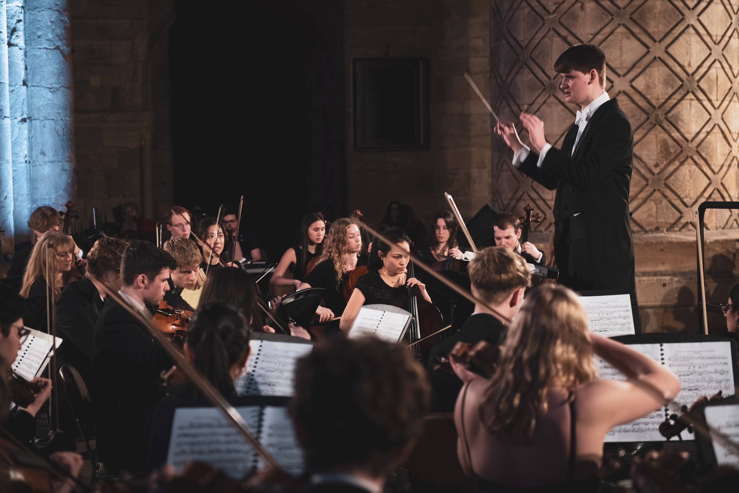 A conductor leading an orchestra of string musicians performing inside a church or historical building.
