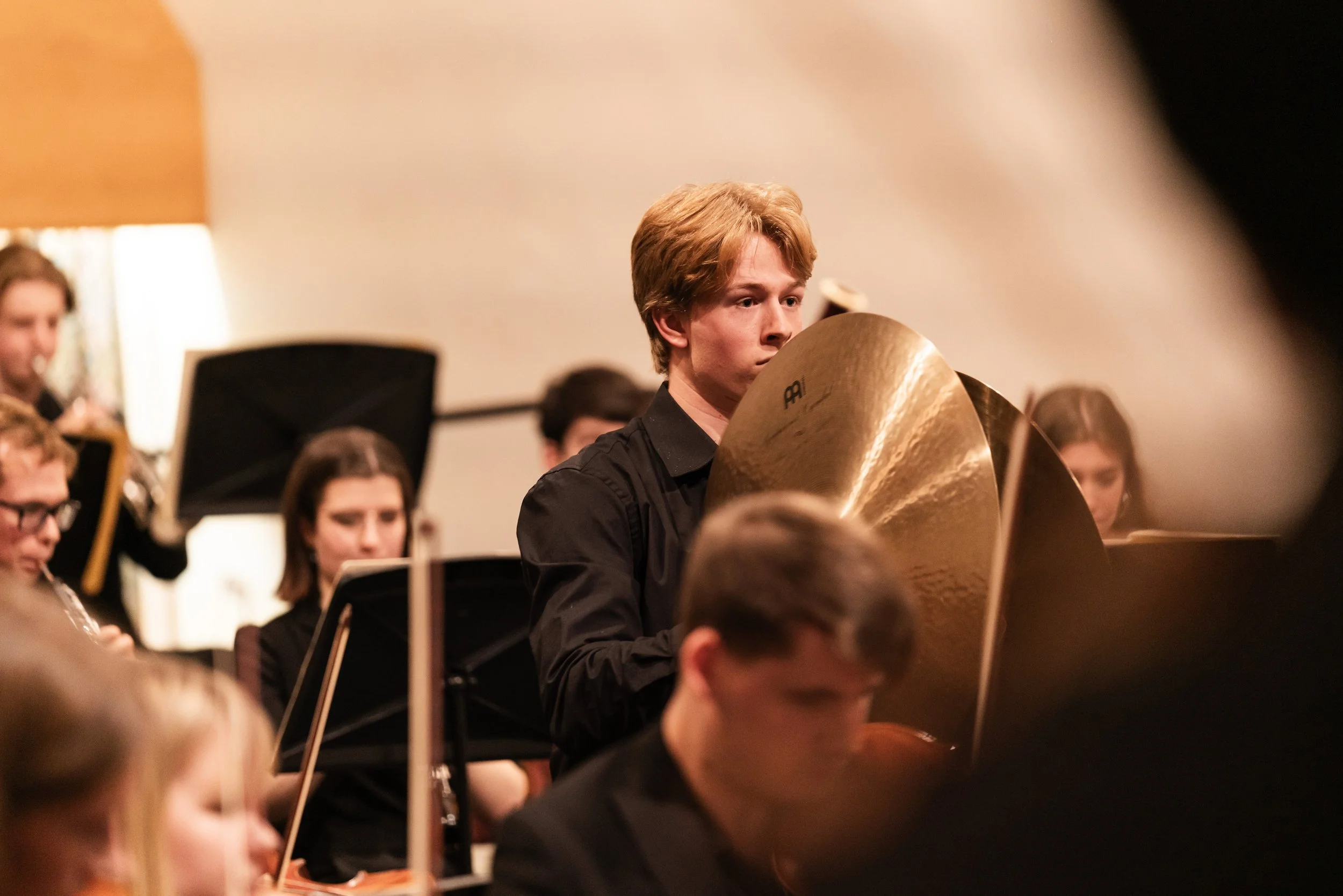 Young musician with light brown hair playing large brass cymbals during a concert, surrounded by other orchestra members with music stands and sheet music.
