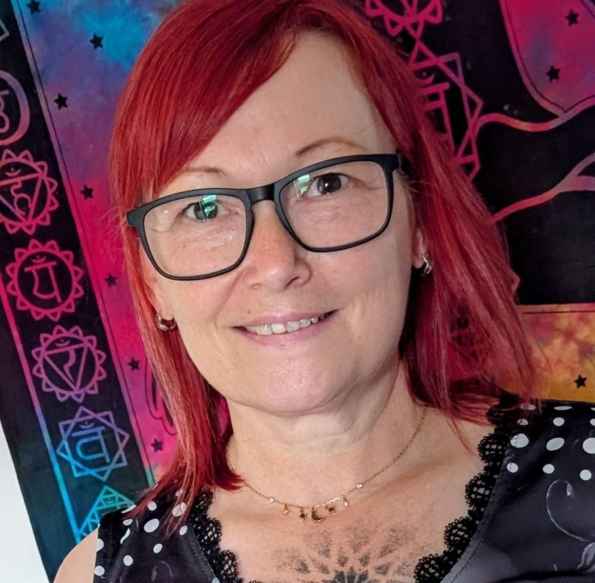 A woman with red hair, glasses, and earrings, smiling in front of a colorful, tapestry-like background with celestial and geometric designs.