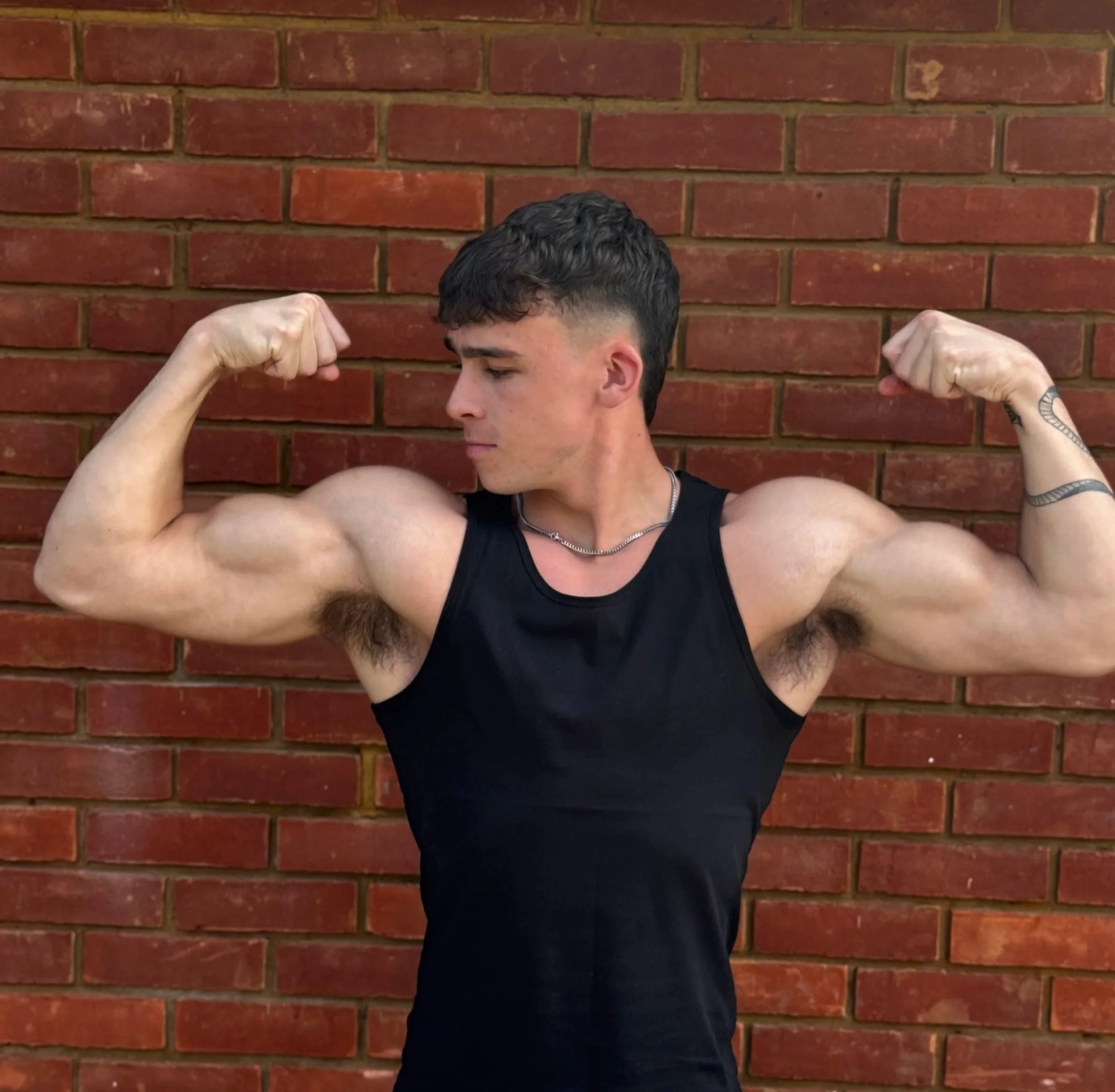 Young man with short dark hair flexing his biceps in front of a brick wall, wearing a black sleeveless shirt and a silver chain necklace.