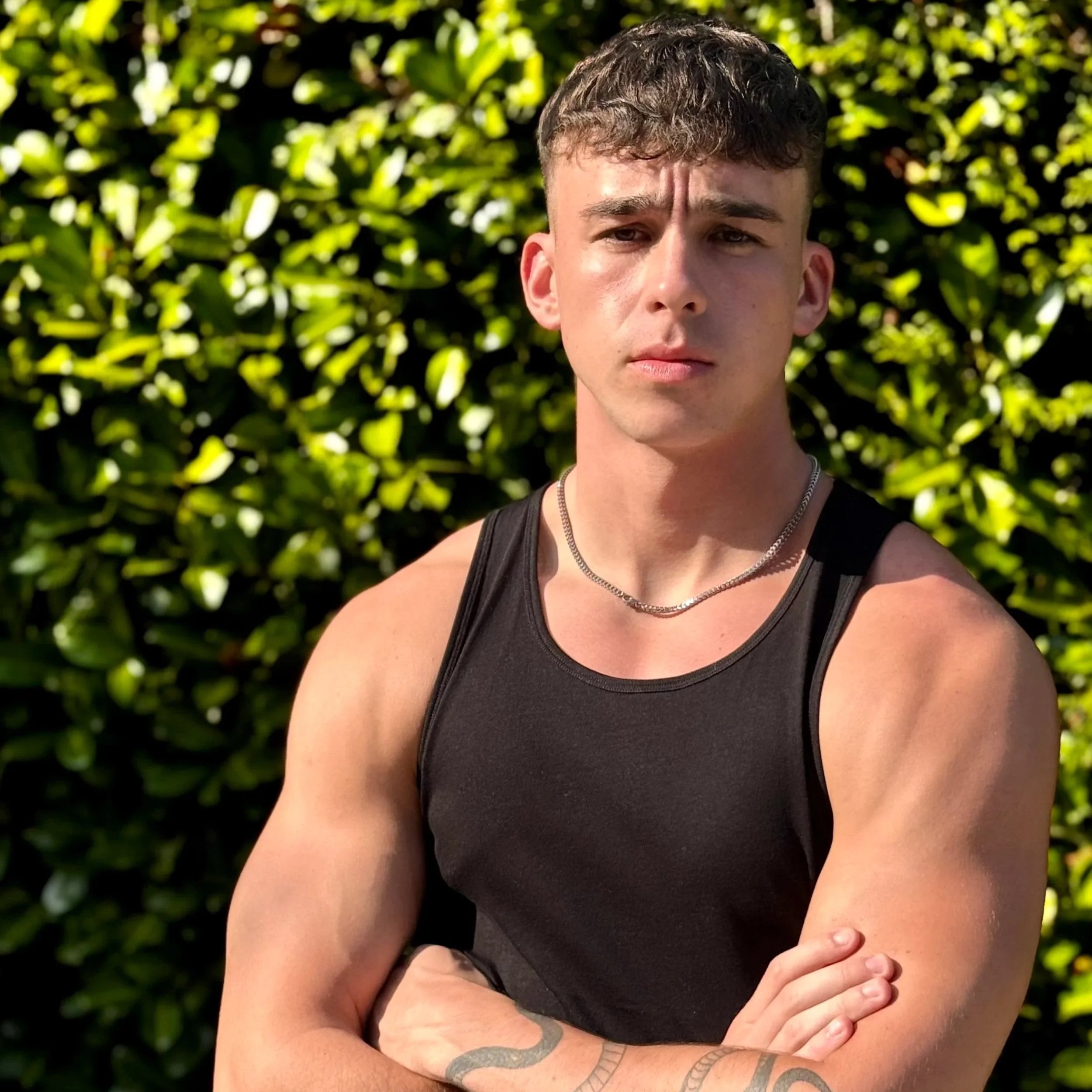 A young man with short brown hair and a serious expression, wearing a black sleeveless tank top and a silver chain necklace, standing outdoors with green foliage in the background.