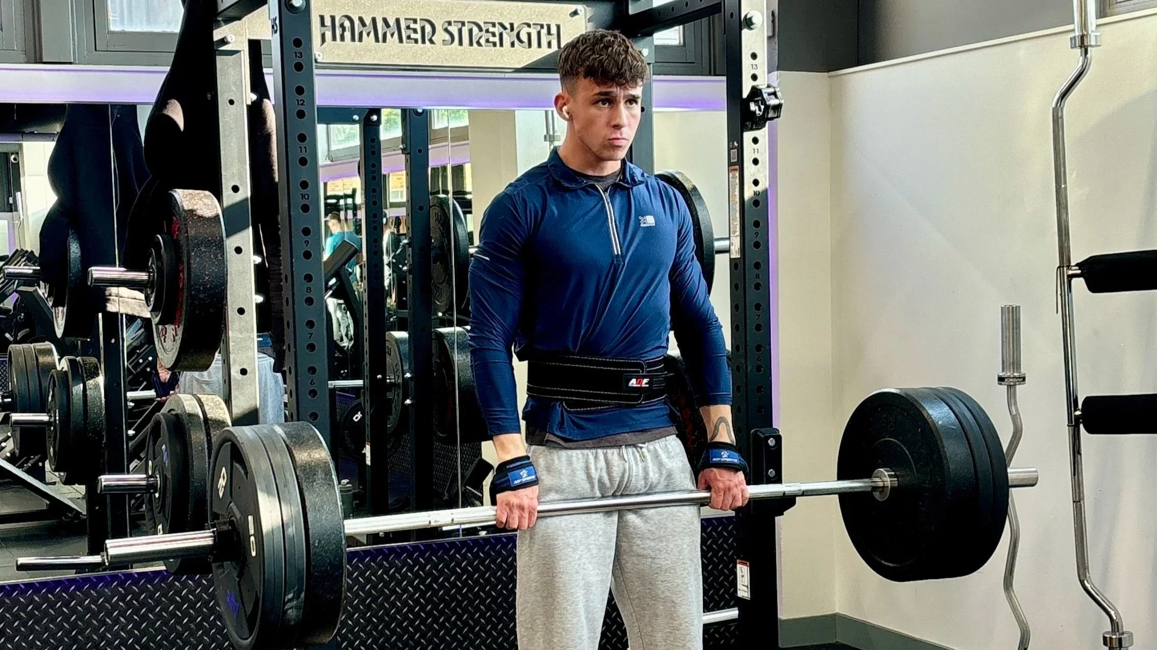Young man in a blue long-sleeve athletic shirt, gray sweatpants, and lifting gloves, performing a deadlift exercise with a barbell loaded with weight plates at a gym. He is standing in front of a power rack with other gym equipment visible in the background.