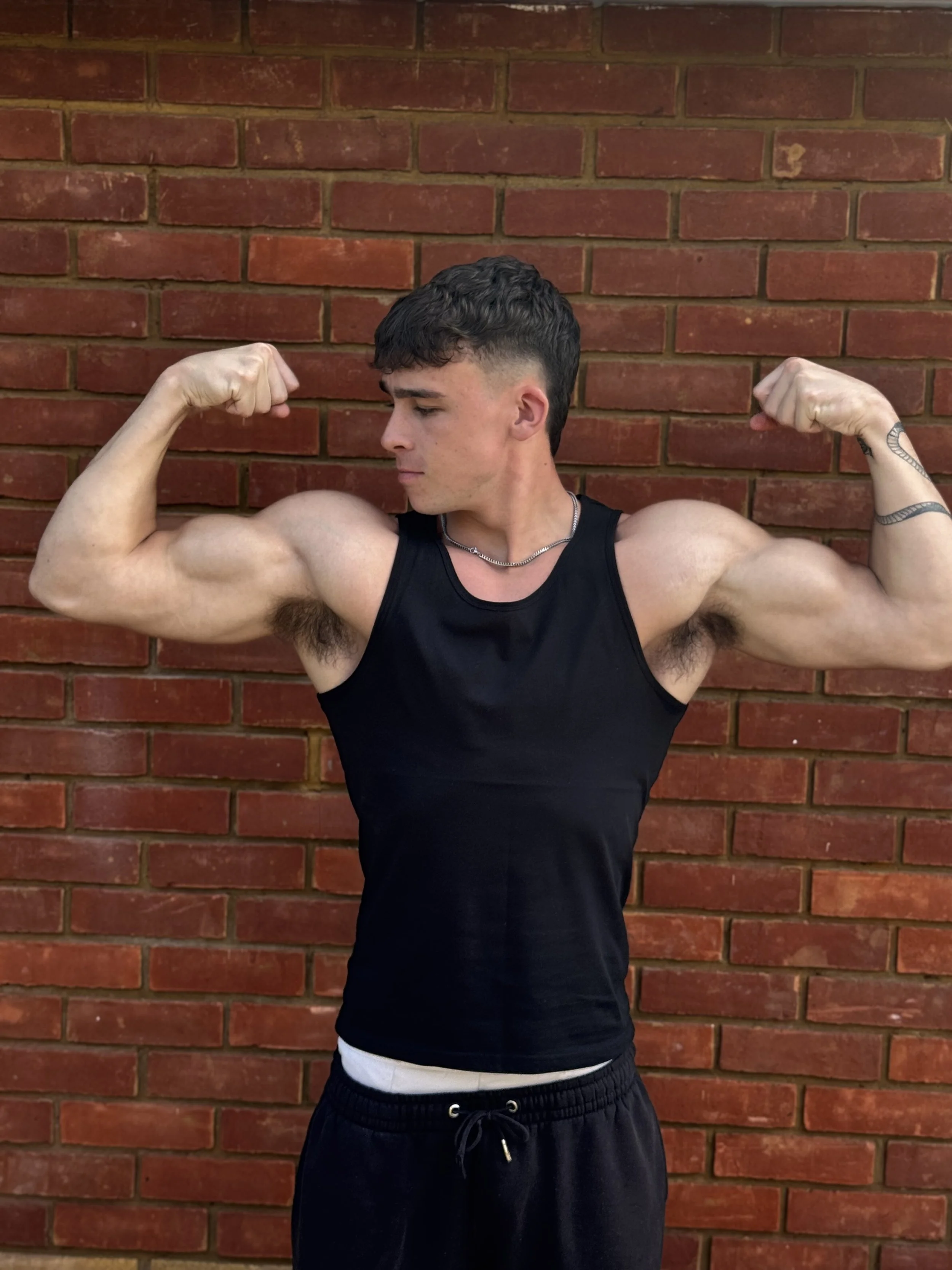 Young man flexing his biceps in front of a brick wall, wearing a black tank top and black shorts.