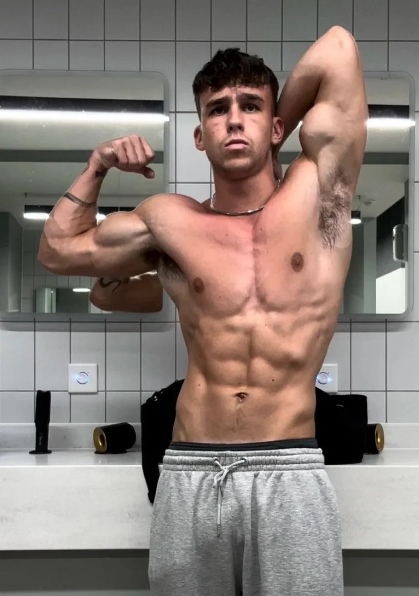 A shirtless young man flexing his arm in front of a bathroom mirror, showing his muscular physique with defined abs and arm muscles.