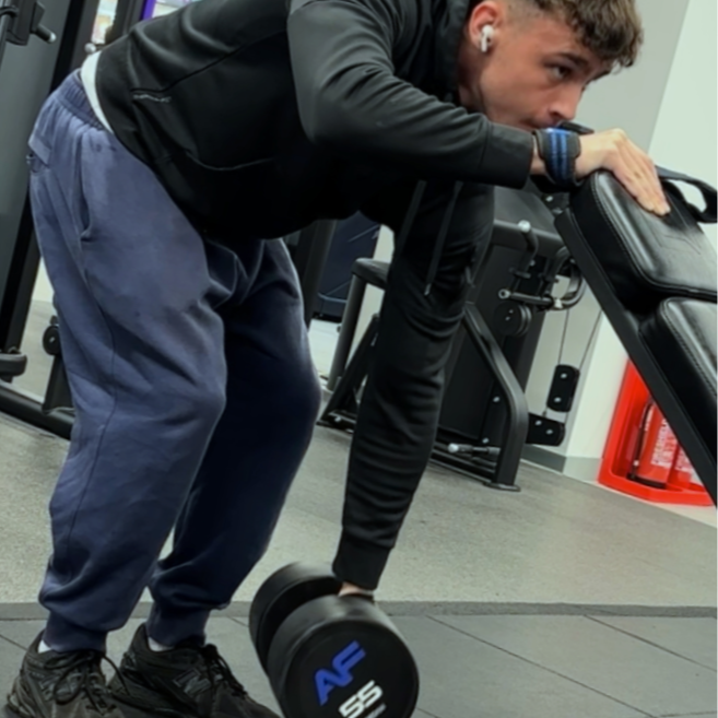 Young man at the gym, bending over to lift a dumbbell with one hand while supporting his body with the other hand on equipment.