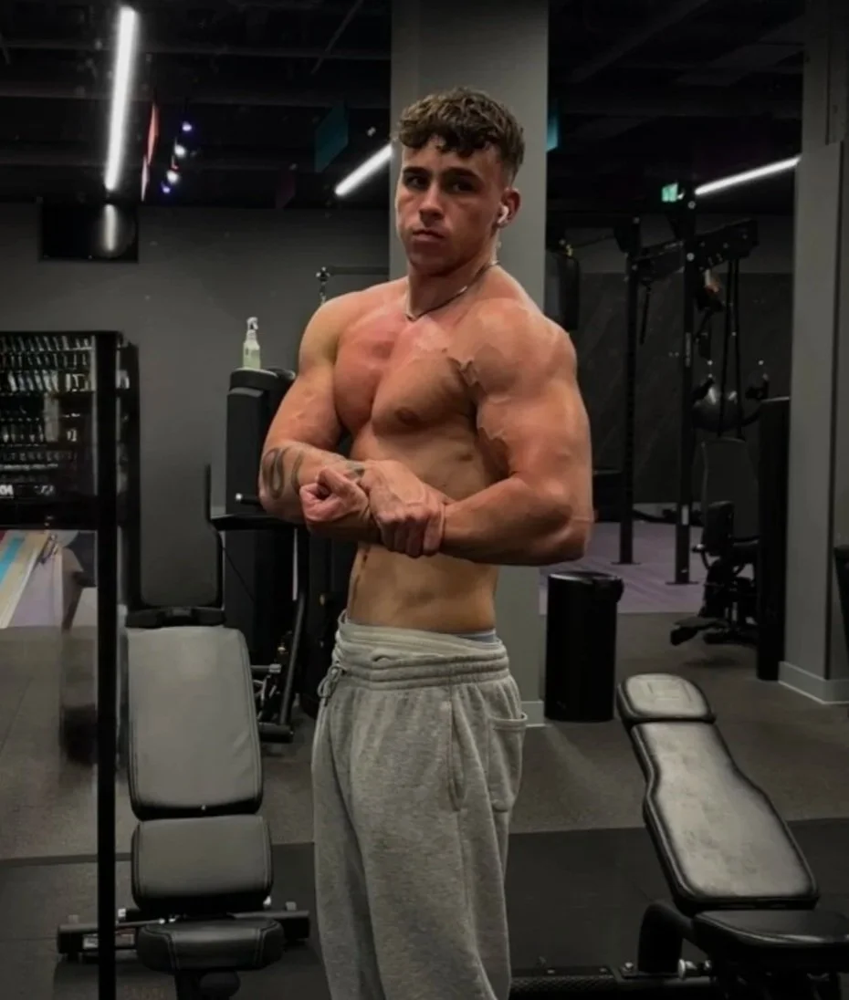 Young man with muscular build, shirtless, flexing his biceps in a gym, wearing gray sweatpants, with gym equipment and mirrors in the background.