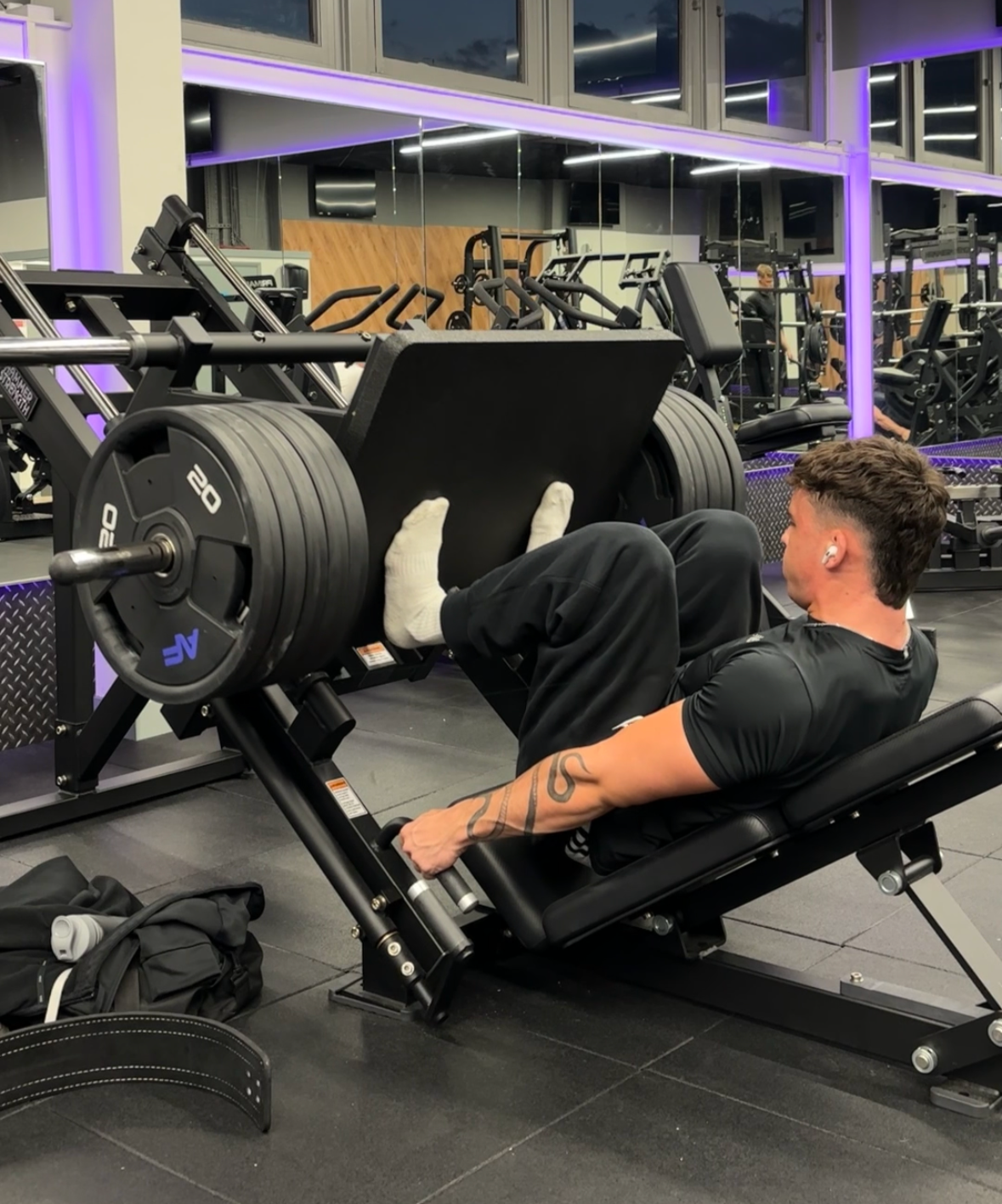 A man is lying on a leg press machine at a gym, lifting large weighted plates with his legs.