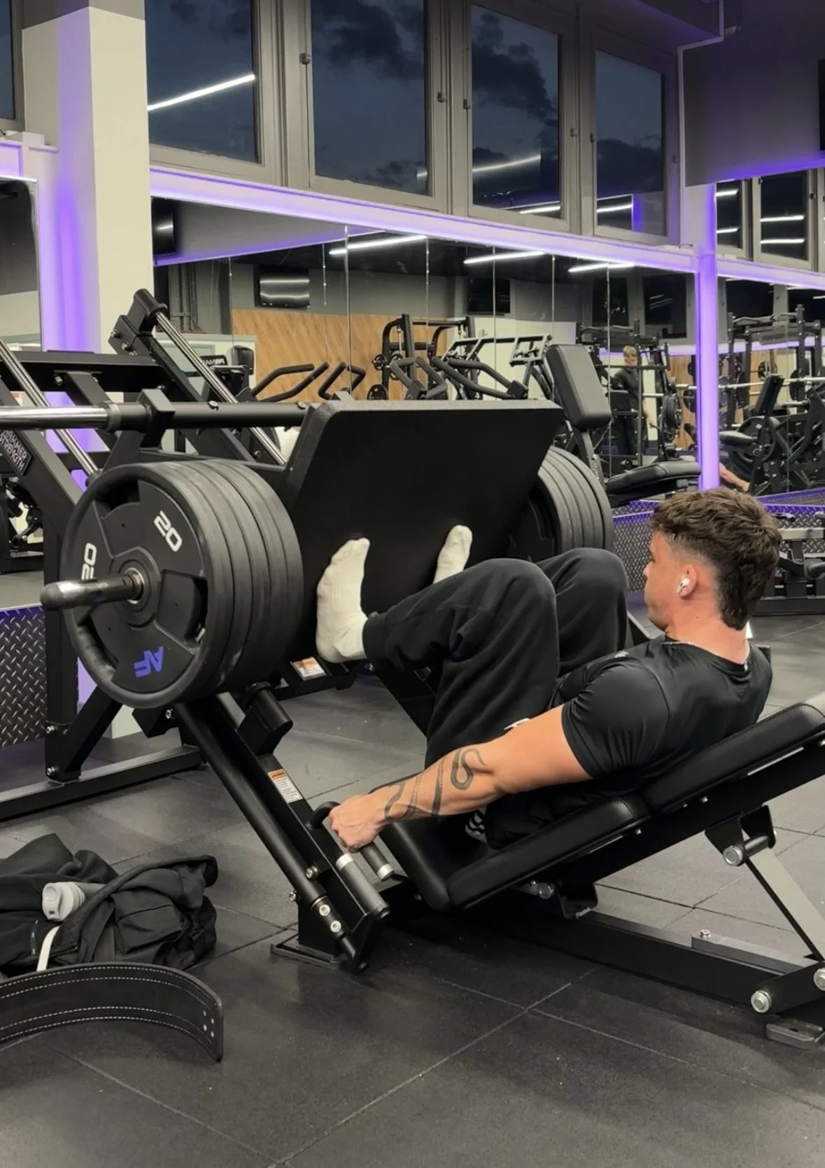 A man in a gym lifting heavy weights on a leg press machine. The gym has modern equipment and purple lighting accents.