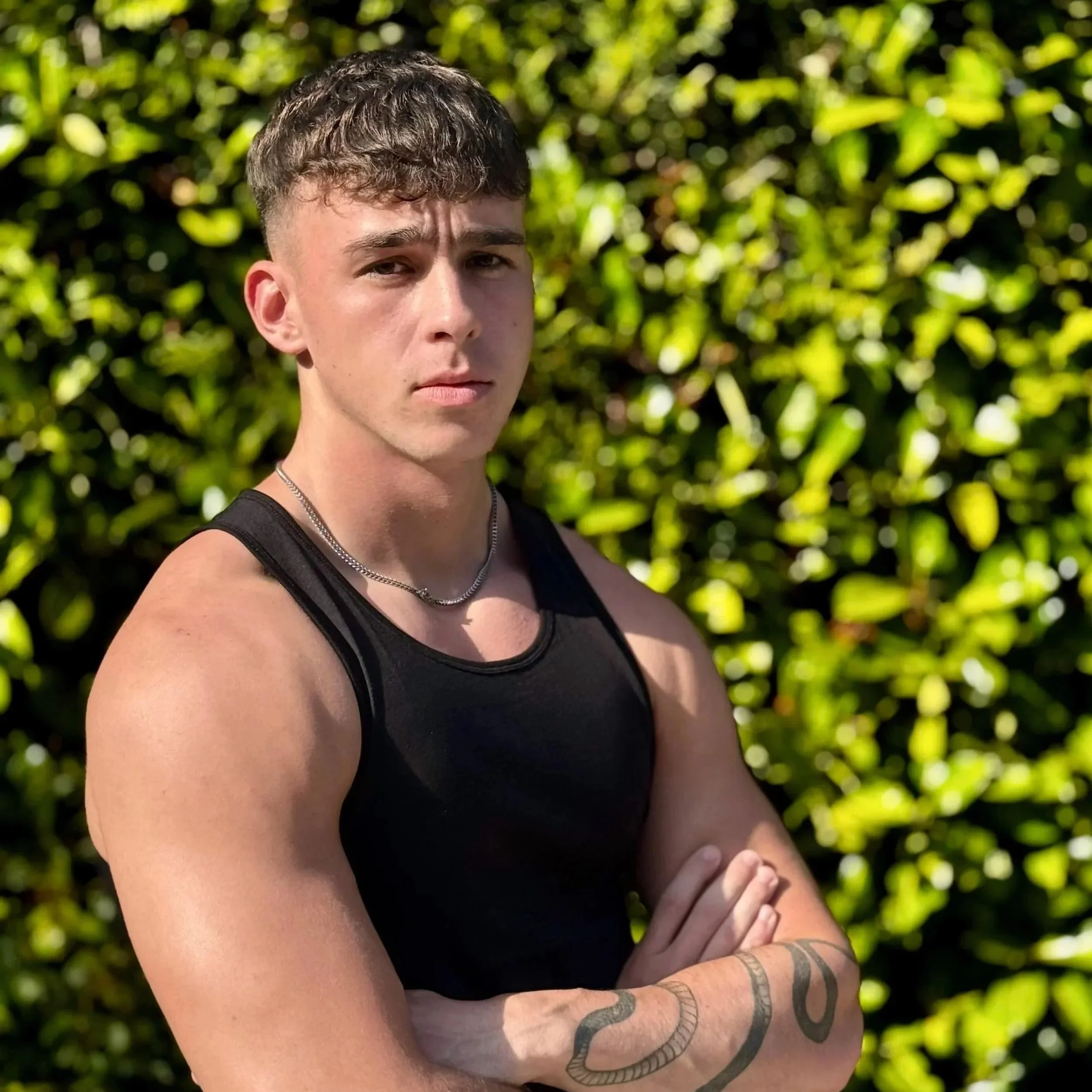 Young man with light skin, short brown hair, and wearing a black tank top, standing outdoors with green leafy bushes in the background, crossing his arms and looking serious.
