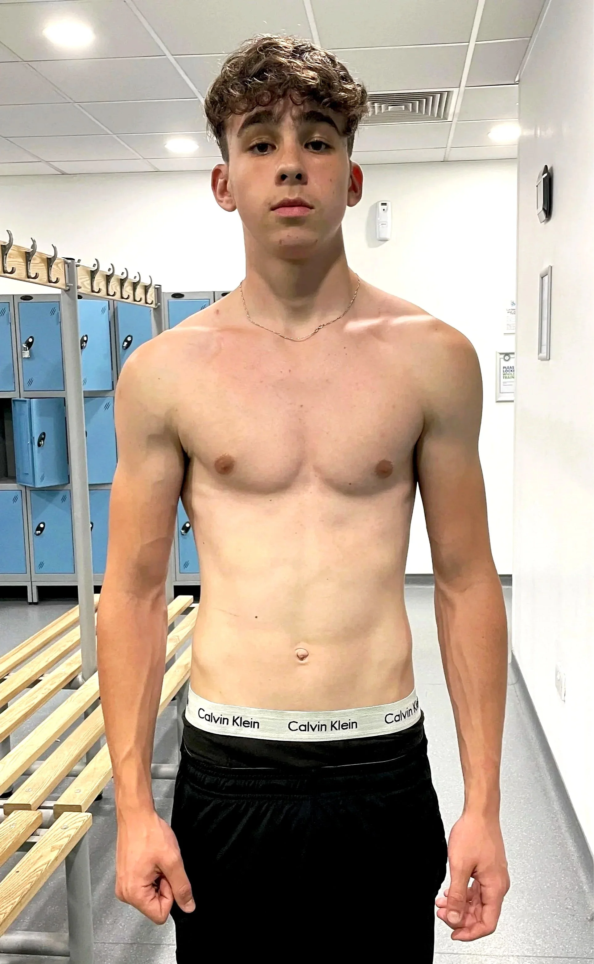 A young shirtless man standing in a locker room with blue lockers in the background.