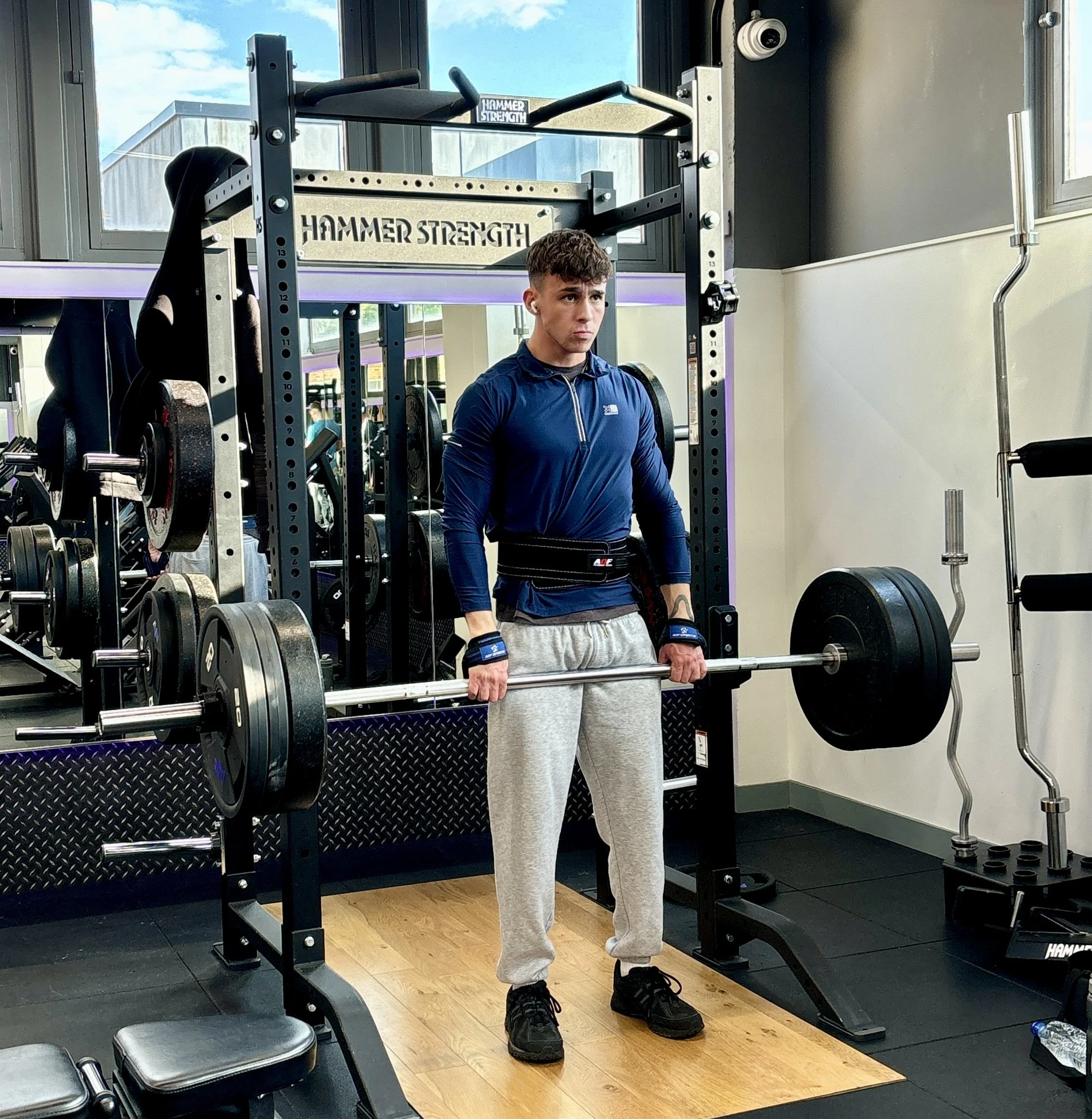 A young man lifting a barbell with heavy weights in a gym, standing on a wooden platform surrounded by gym equipment and mirrors.