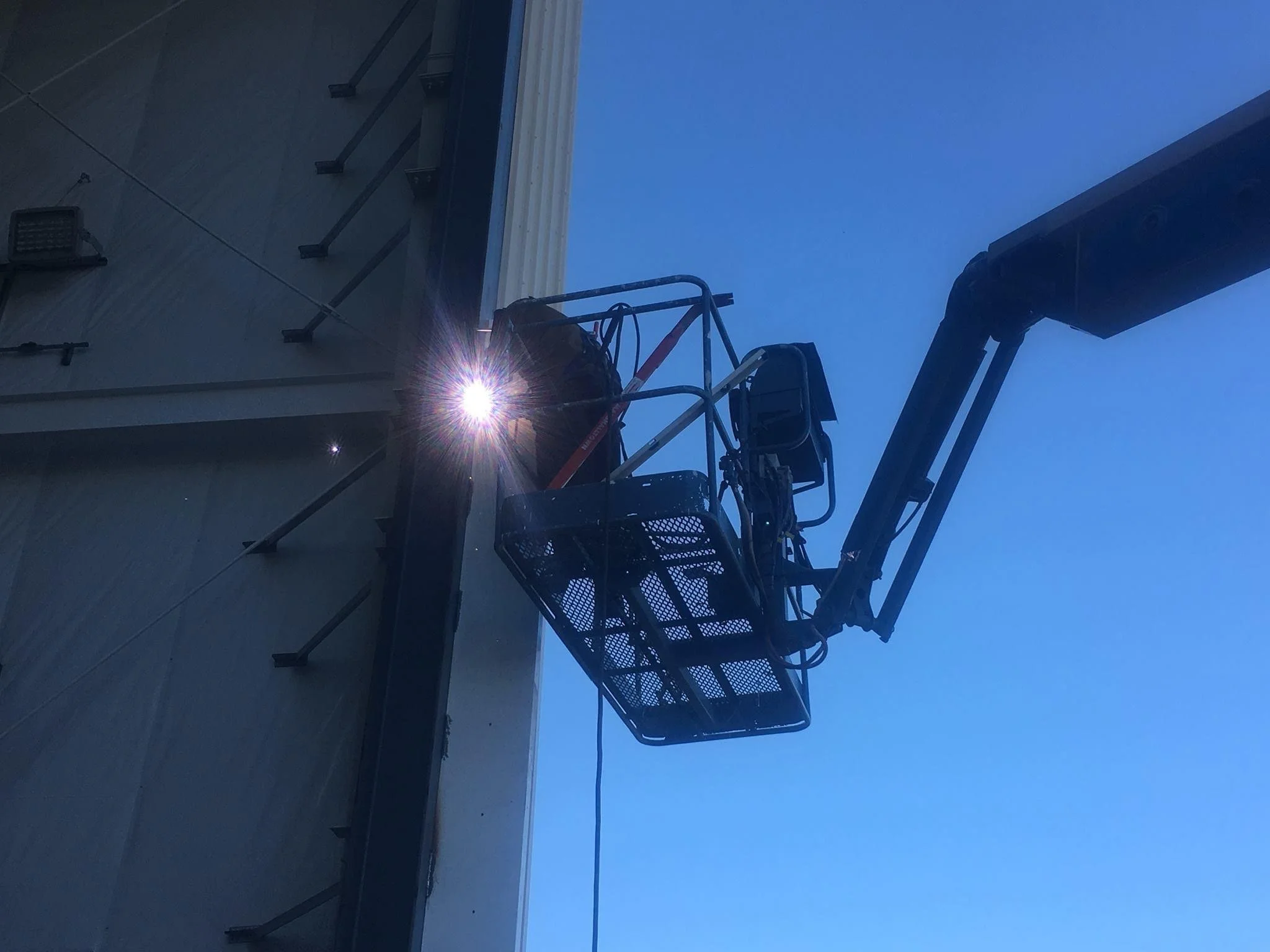 A worker on a scissor lift welding on the exterior of a building, with the welding sparks visible against a clear blue sky.