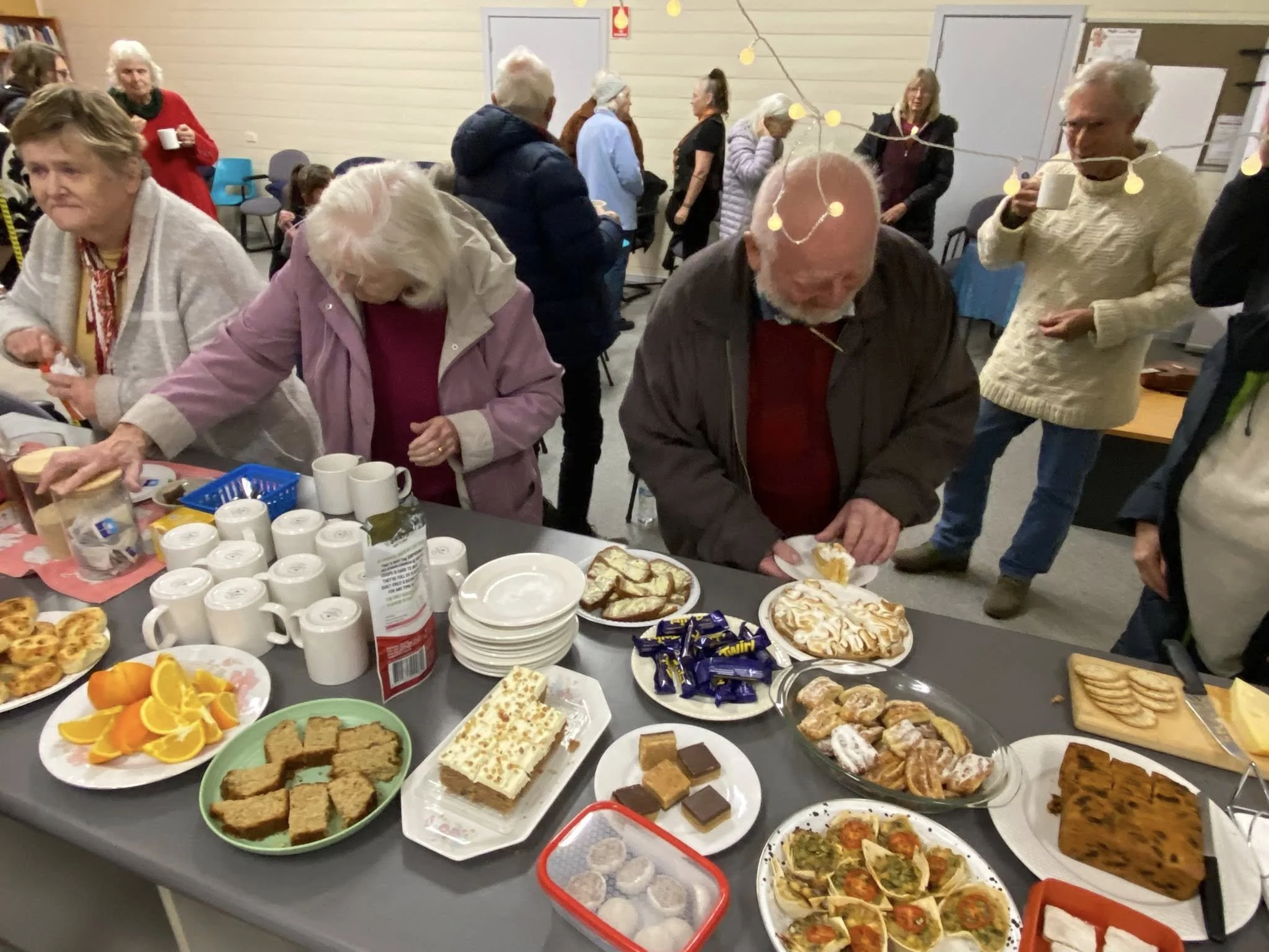 A group of elderly people at a gathering around a table with various desserts and snacks, including plates of cake, cookies, and fruit, in a room decorated with string lights.