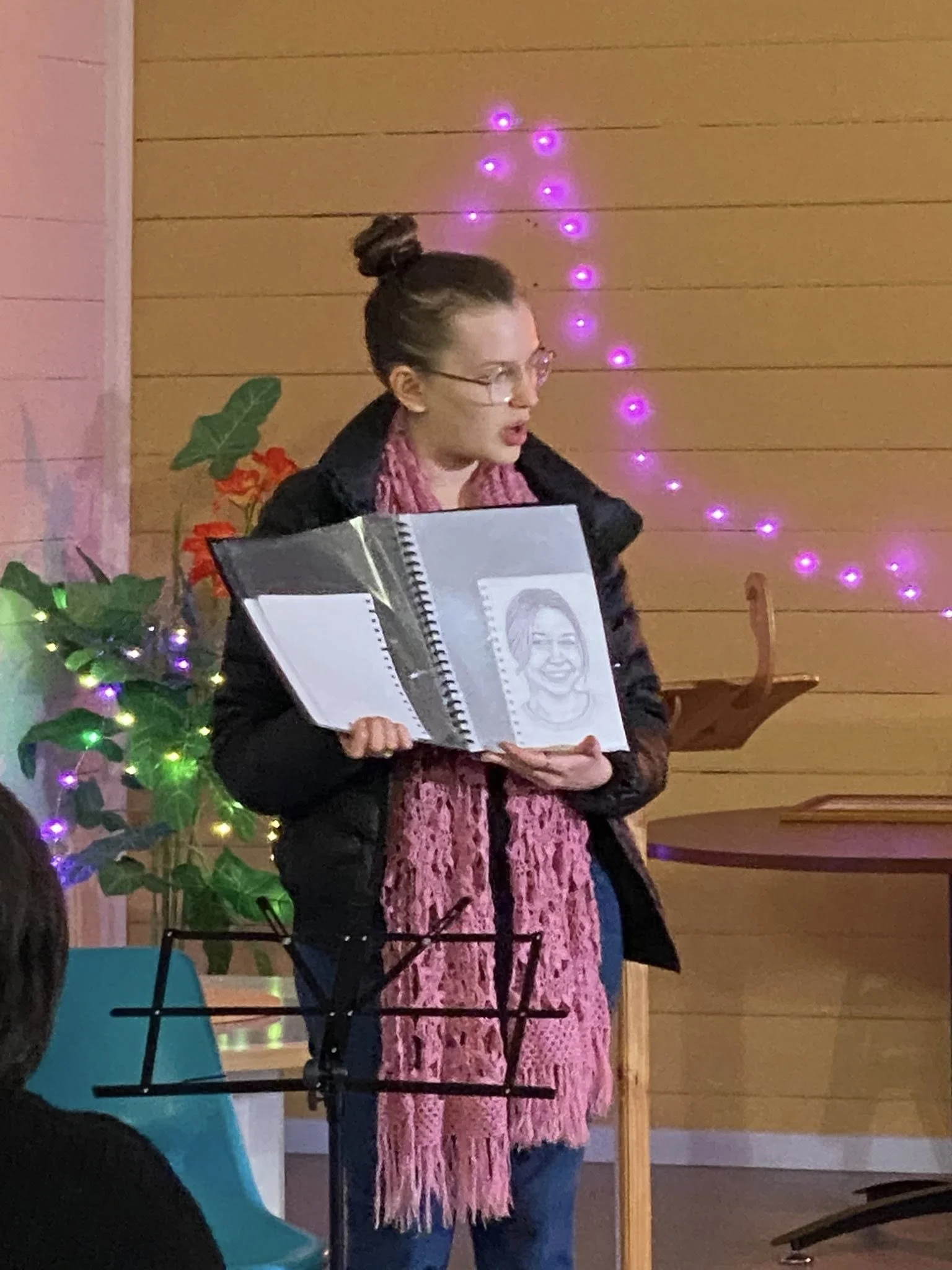 A woman wearing glasses, dressed in a black jacket and a pink scarf, is holding a large sketchbook open and presenting a drawing of a smiling woman. The background features a wooden wall decorated with colorful string lights and green plants.