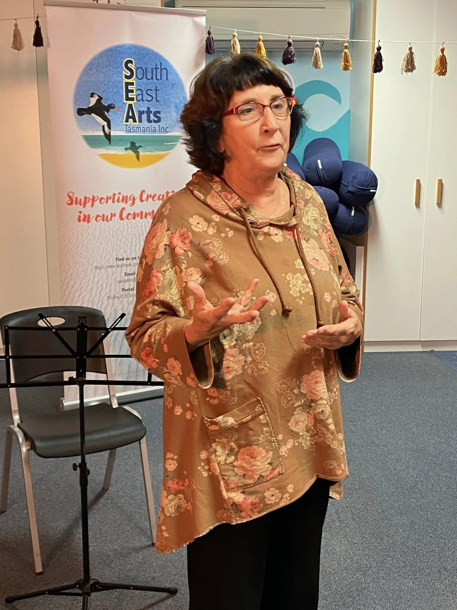 A woman wearing glasses and a floral patterned, gold-colored jacket speaking indoors, with a South East Arts banner and a chair in the background.