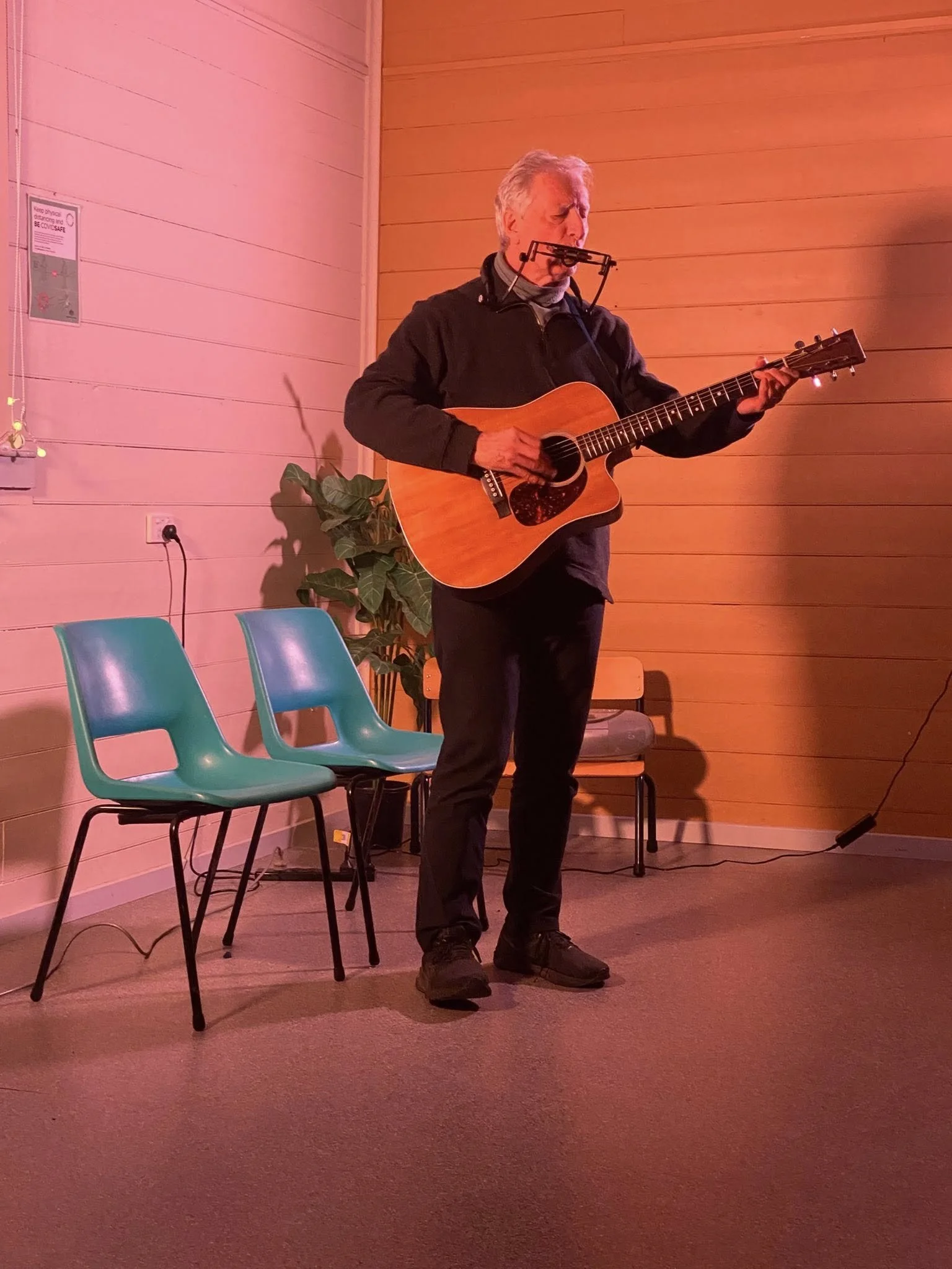 A man playing an acoustic guitar, wearing a black jacket and pants, standing on a stage with wooden walls, next to two teal chairs, with a potted plant and chair in the background, during a live performance.