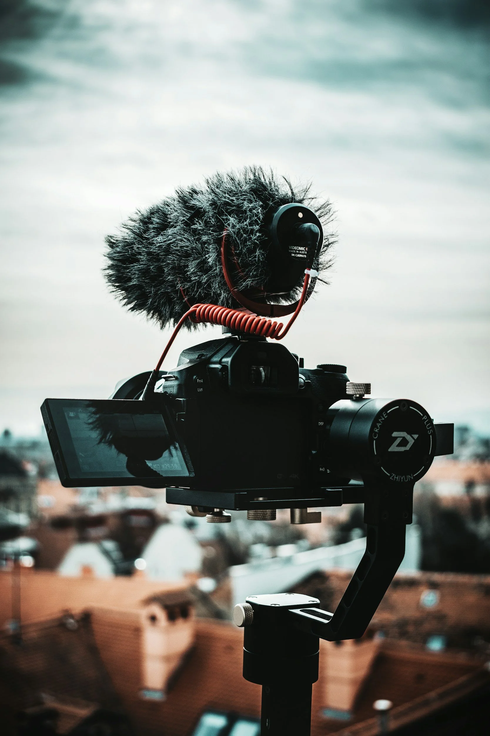 Video camera with a microphone mounted on top, set outdoors with a blurred cityscape background, under a cloudy sky.