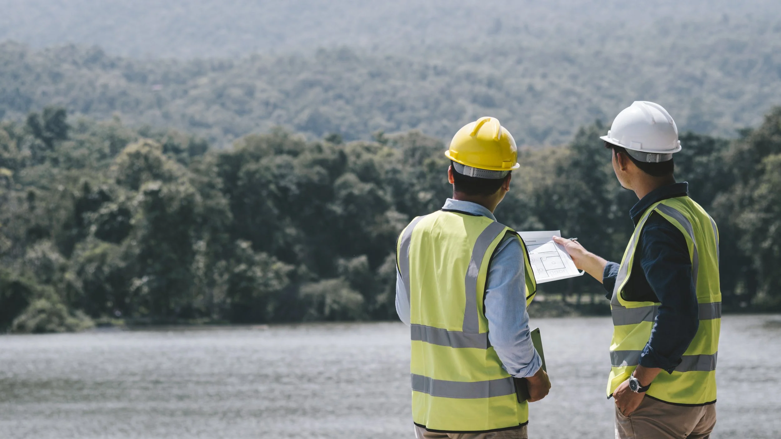 Two construction workers wearing yellow and white safety helmets and yellow reflective vests stand near a body of water with a wooded hill in the background. One worker is holding a tablet, and the other is holding a document, both looking at the document.