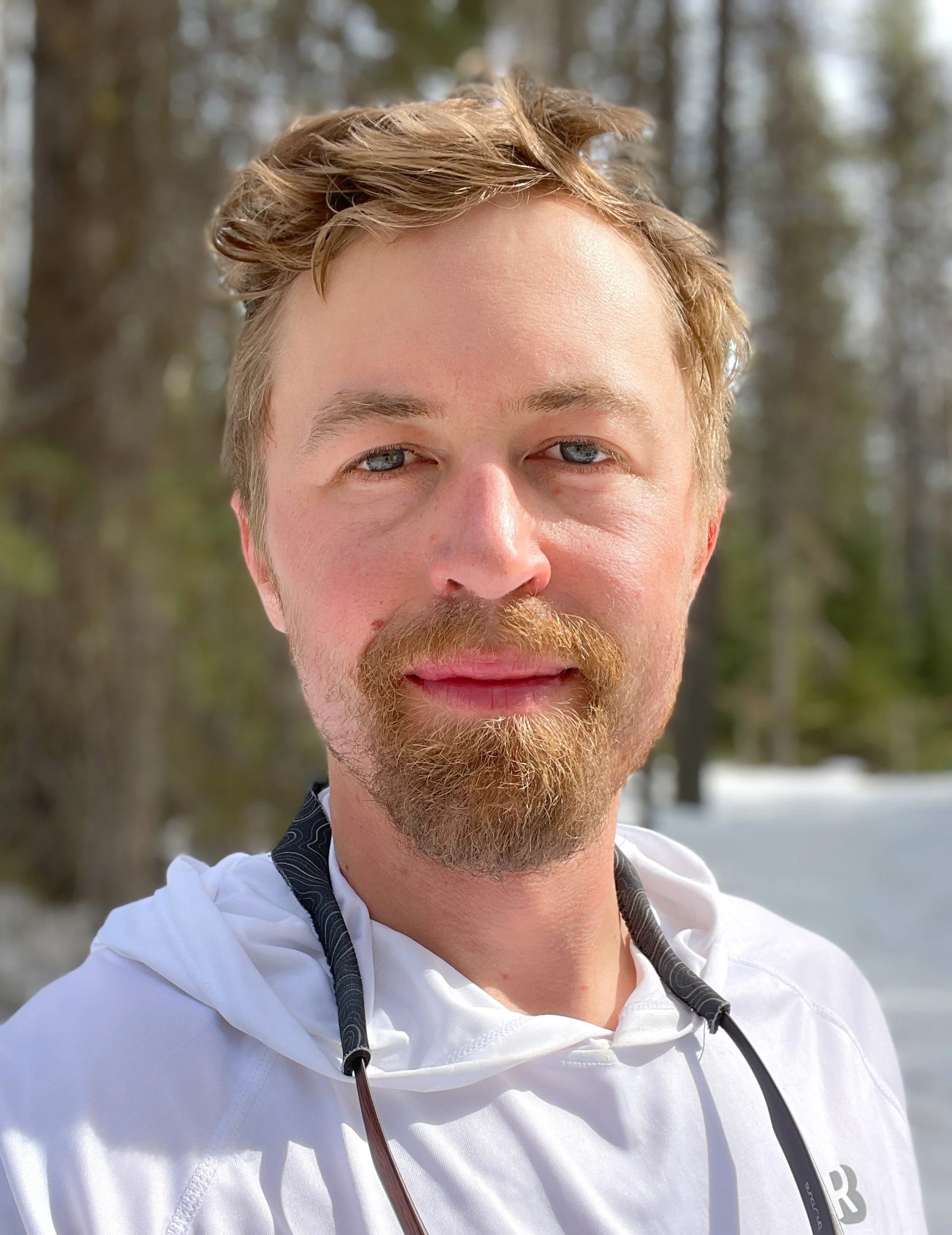 A close-up selfie of a man with light brown hair, a beard, and blue eyes, wearing a white jacket outdoors in a snowy forested area.
