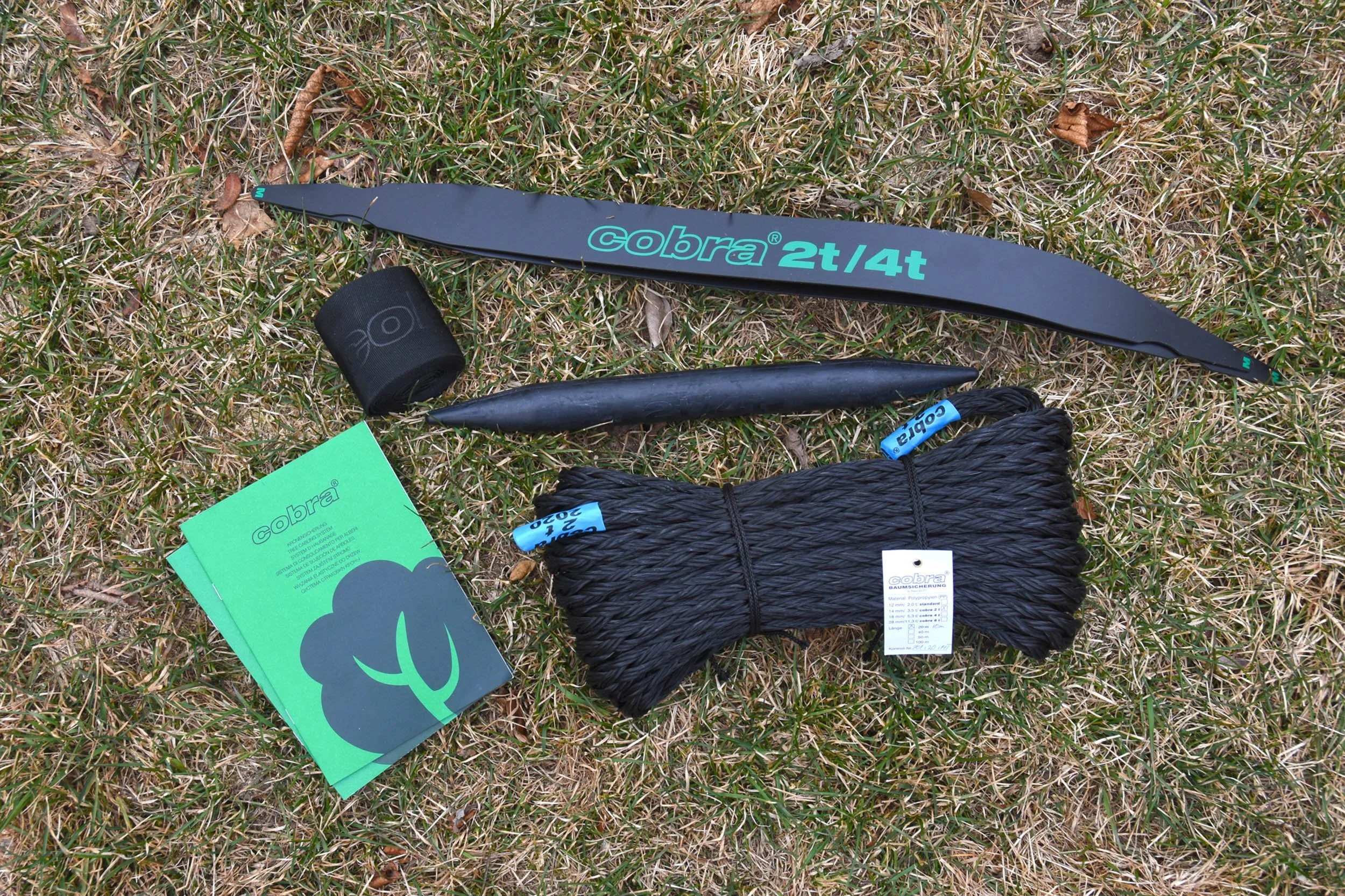 Climbing gear including a black and blue Cobra rope, a black chalk bag, a black holds, a roll of athletic tape, and several green instruction booklets on a grassy ground.