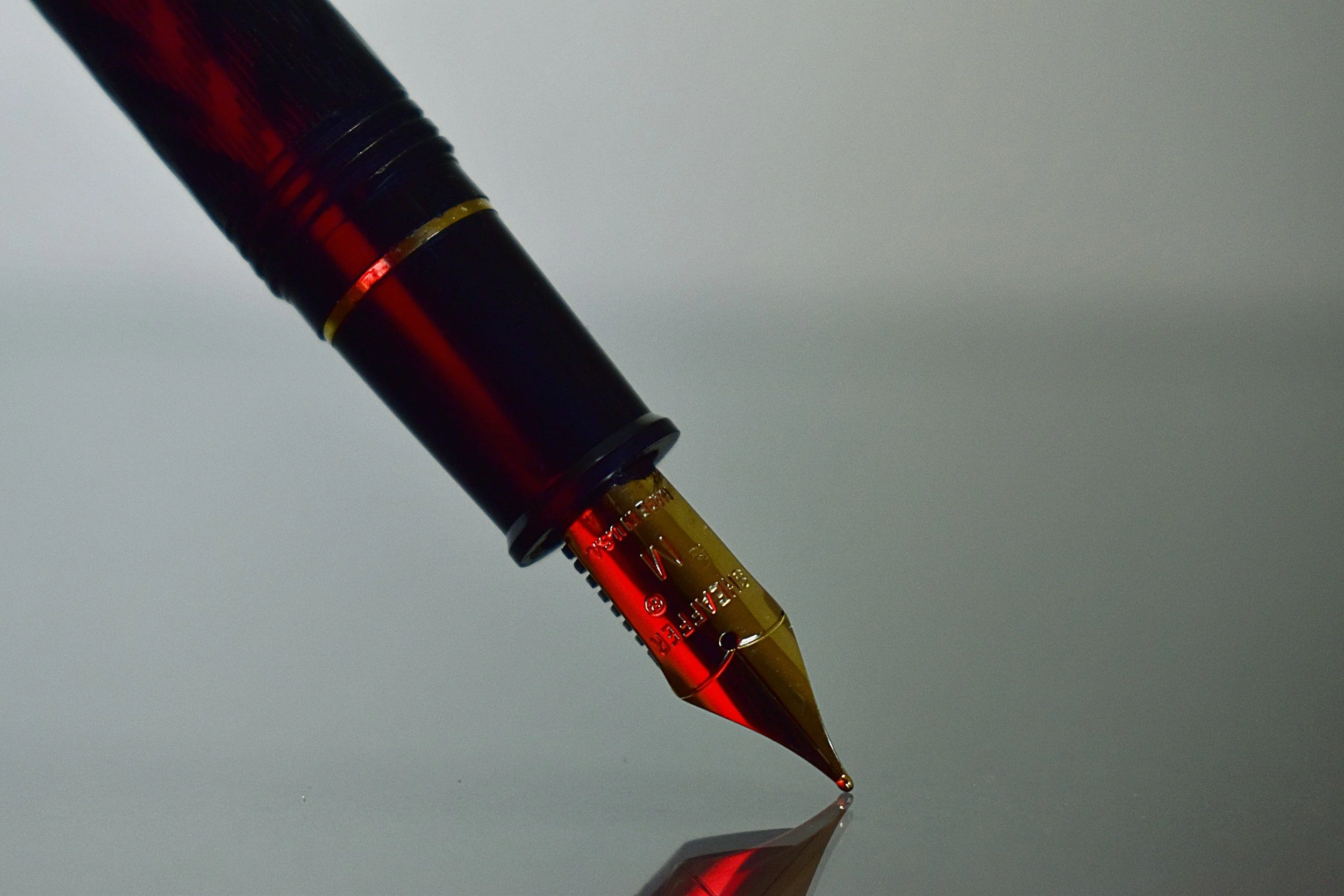 Close-up of a red and black fountain pen tip resting on a reflective surface, with a small ink droplet forming at the tip.